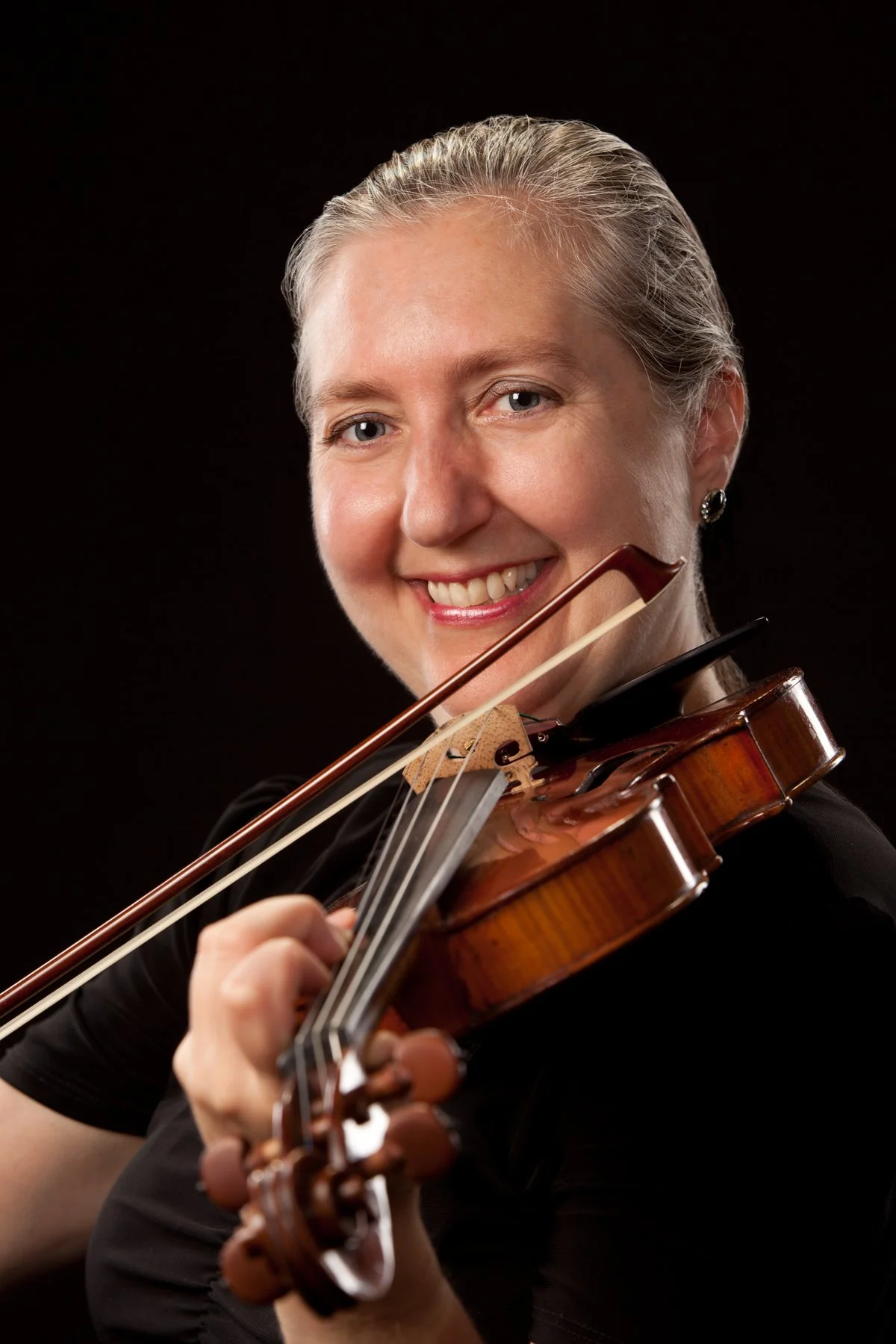 A smiling woman with blonde hair pulled back, playing a violin against a black background.
