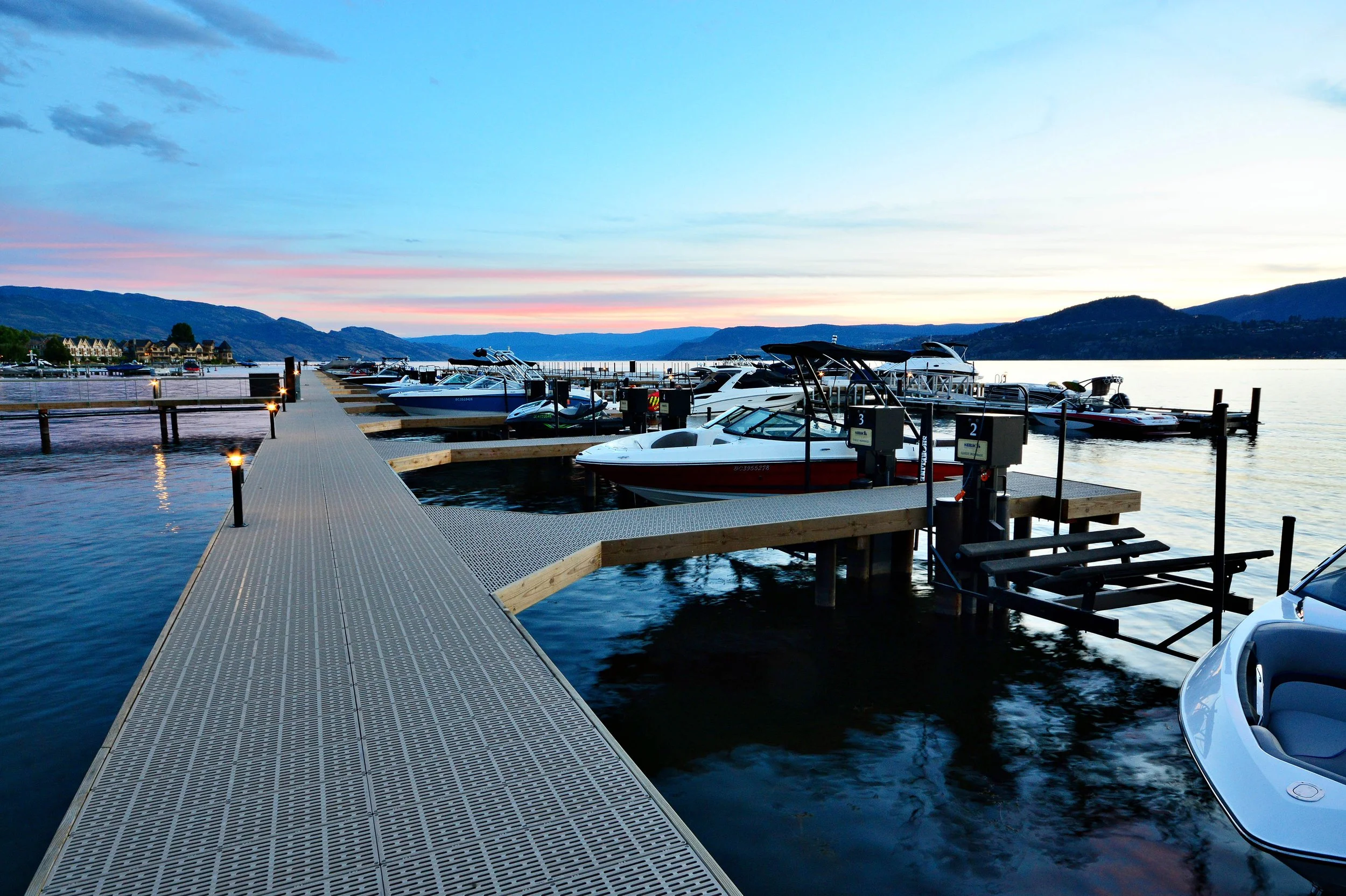 A marina with several boats docked along a pier at sunset, with mountains and a partly cloudy sky in the background.
