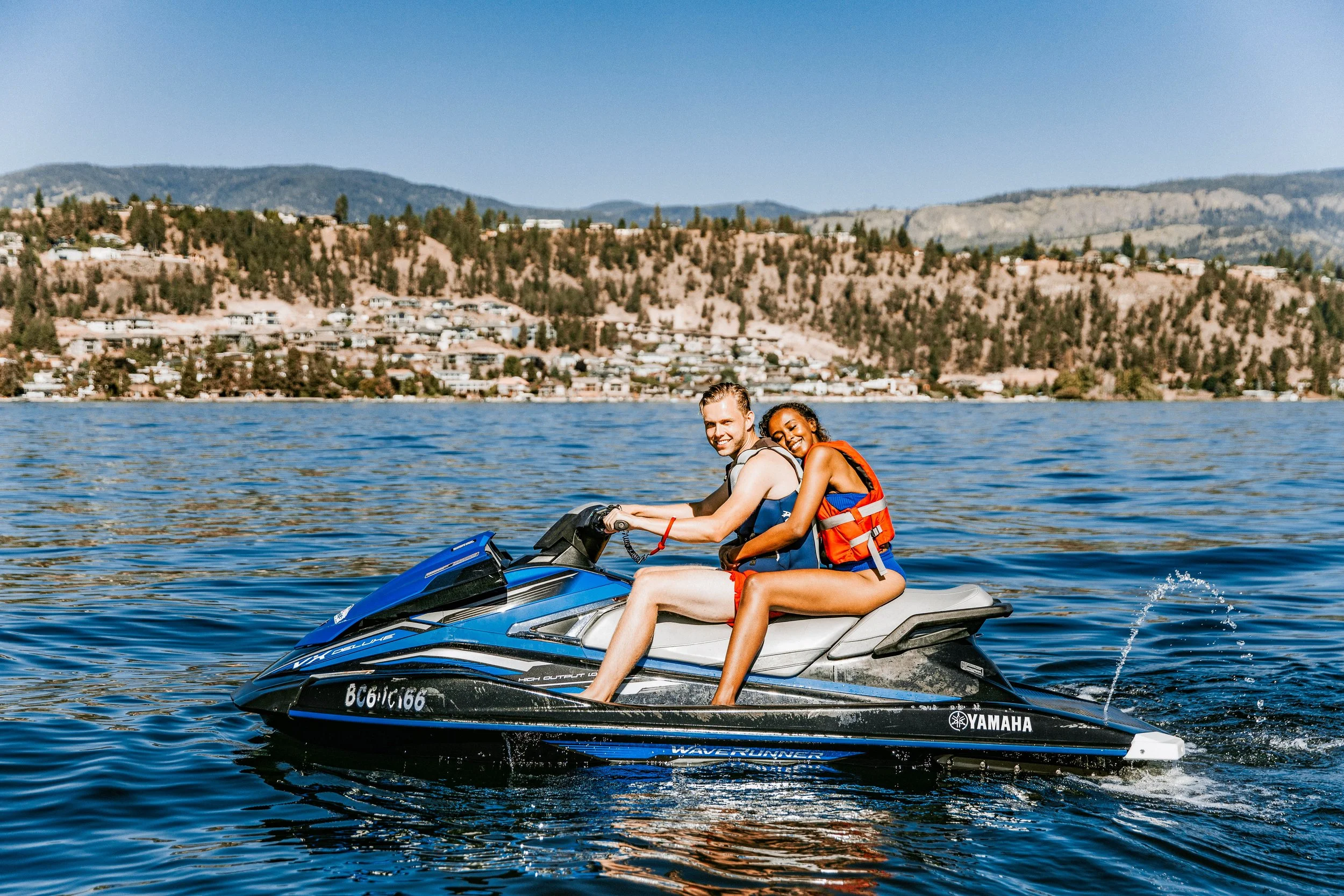 Two people, a man and a woman, riding a Yamaha jet ski on a lake with hills and homes in the background.