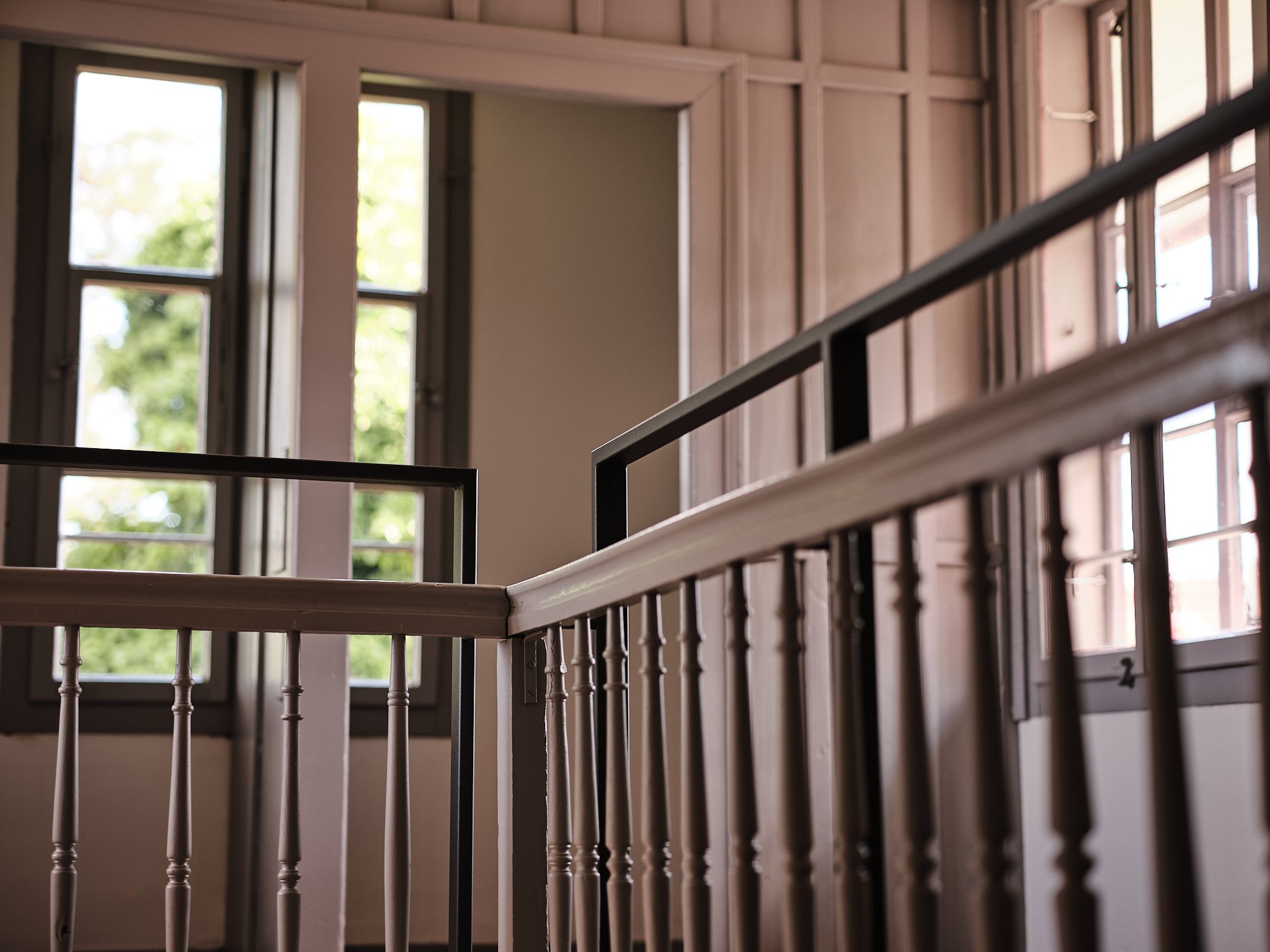 Interior of a building with wooden walls and multiple large windows with natural light, focusing on a staircase railing with vertical balusters.