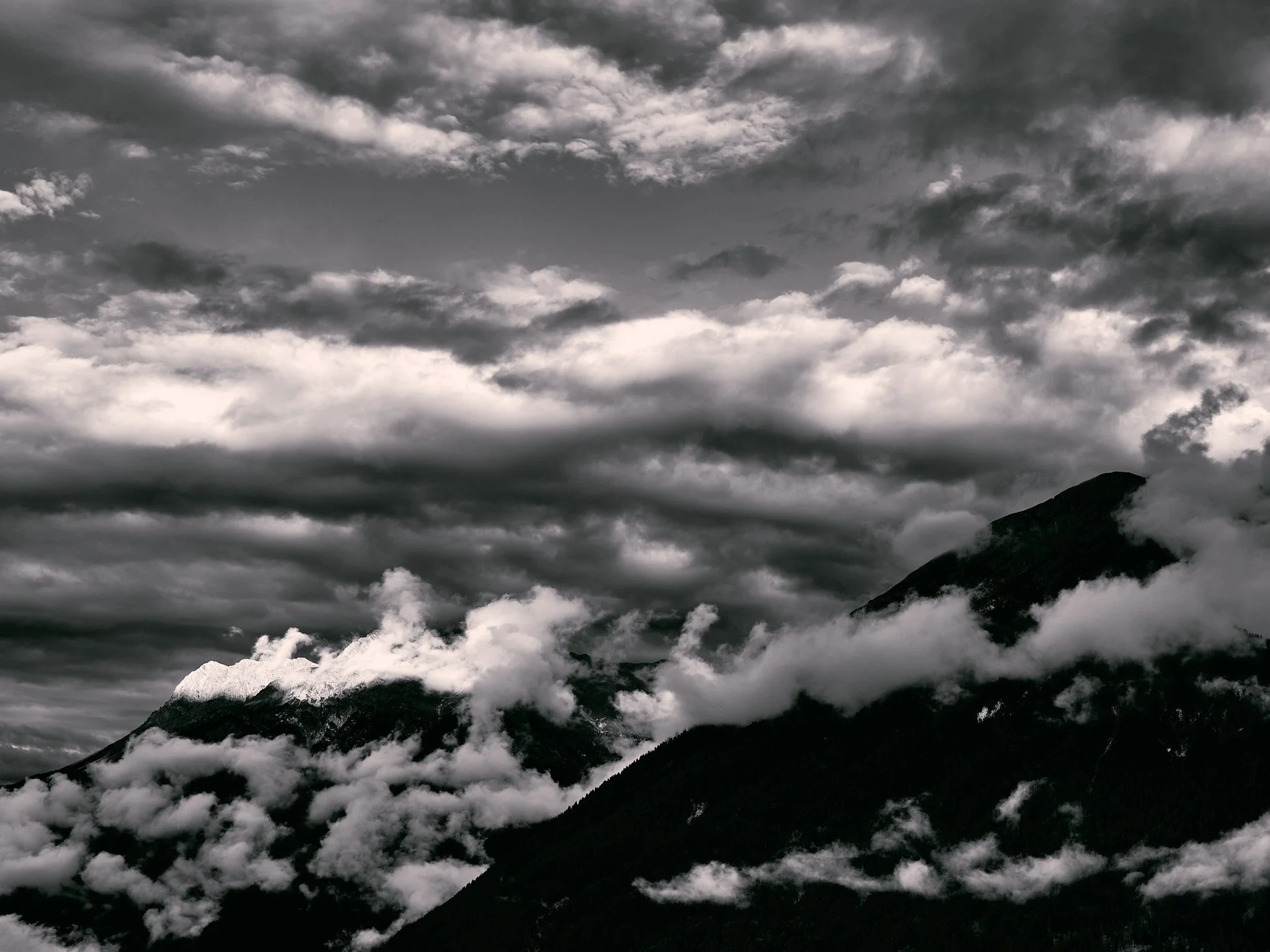 Black and white photo of mountain peaks covered in clouds and mist under a cloudy sky.