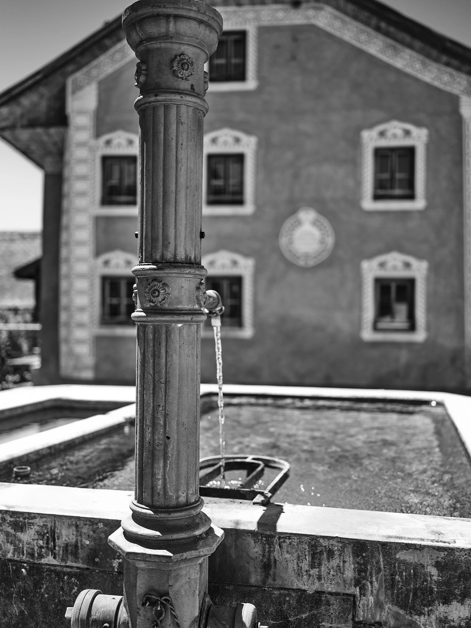 Close-up of an old stone water fountain with a spout of water, in front of a historic building with decorative window frames, in black and white.