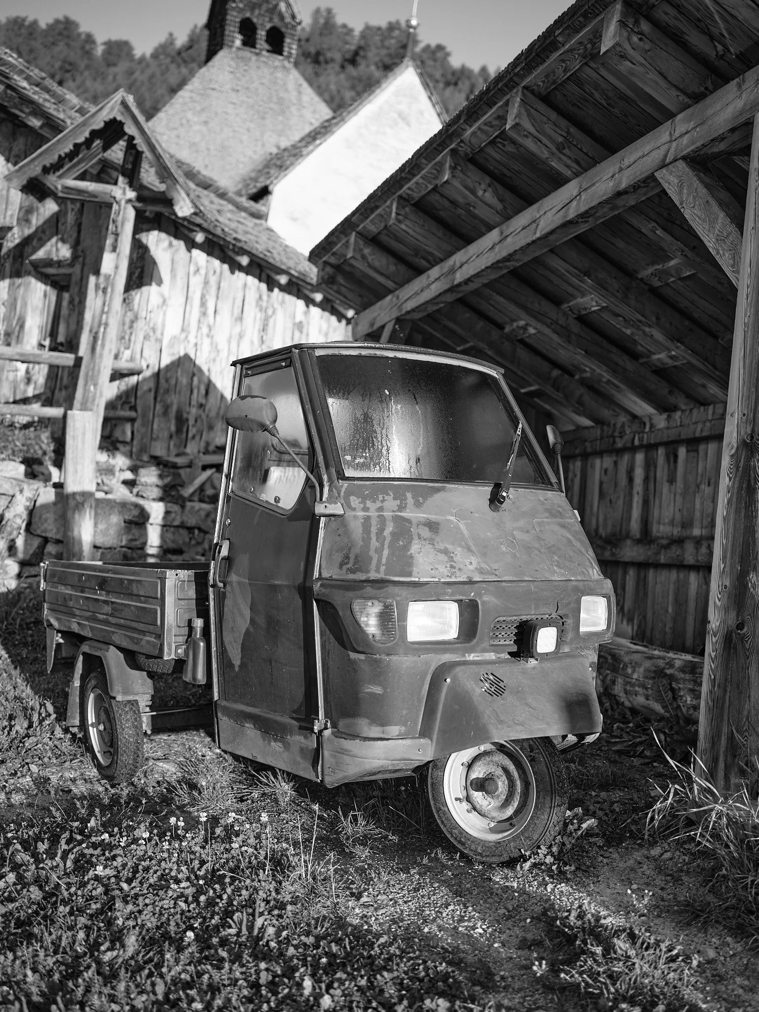 A black and white photo of an old, small, three-wheeled utility vehicle parked under a wooden structure, with a building featuring a dome and tower visible in the background.