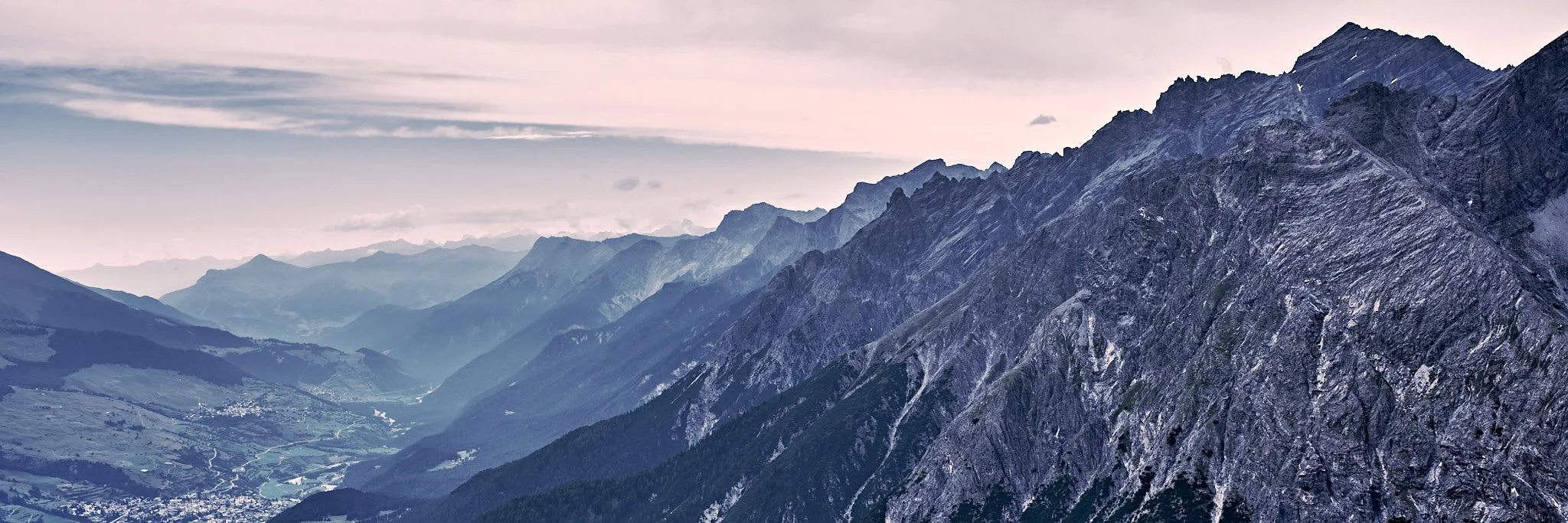 View of a mountain range with rugged peaks and a valley below, under a cloudy sky.