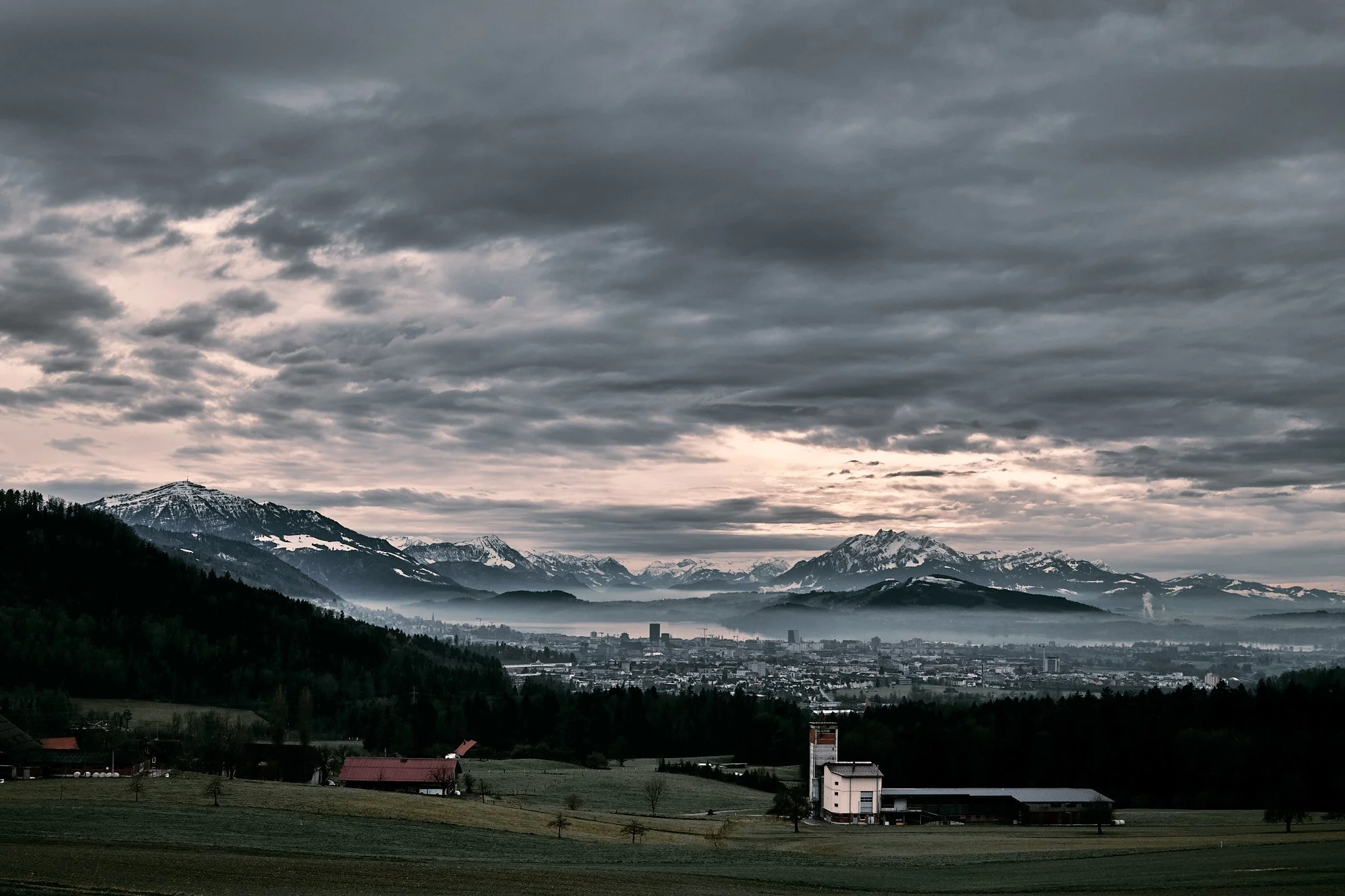 A scenic view of mountains with snow caps, a cloudy sky, and a rural landscape with farm buildings in the foreground.