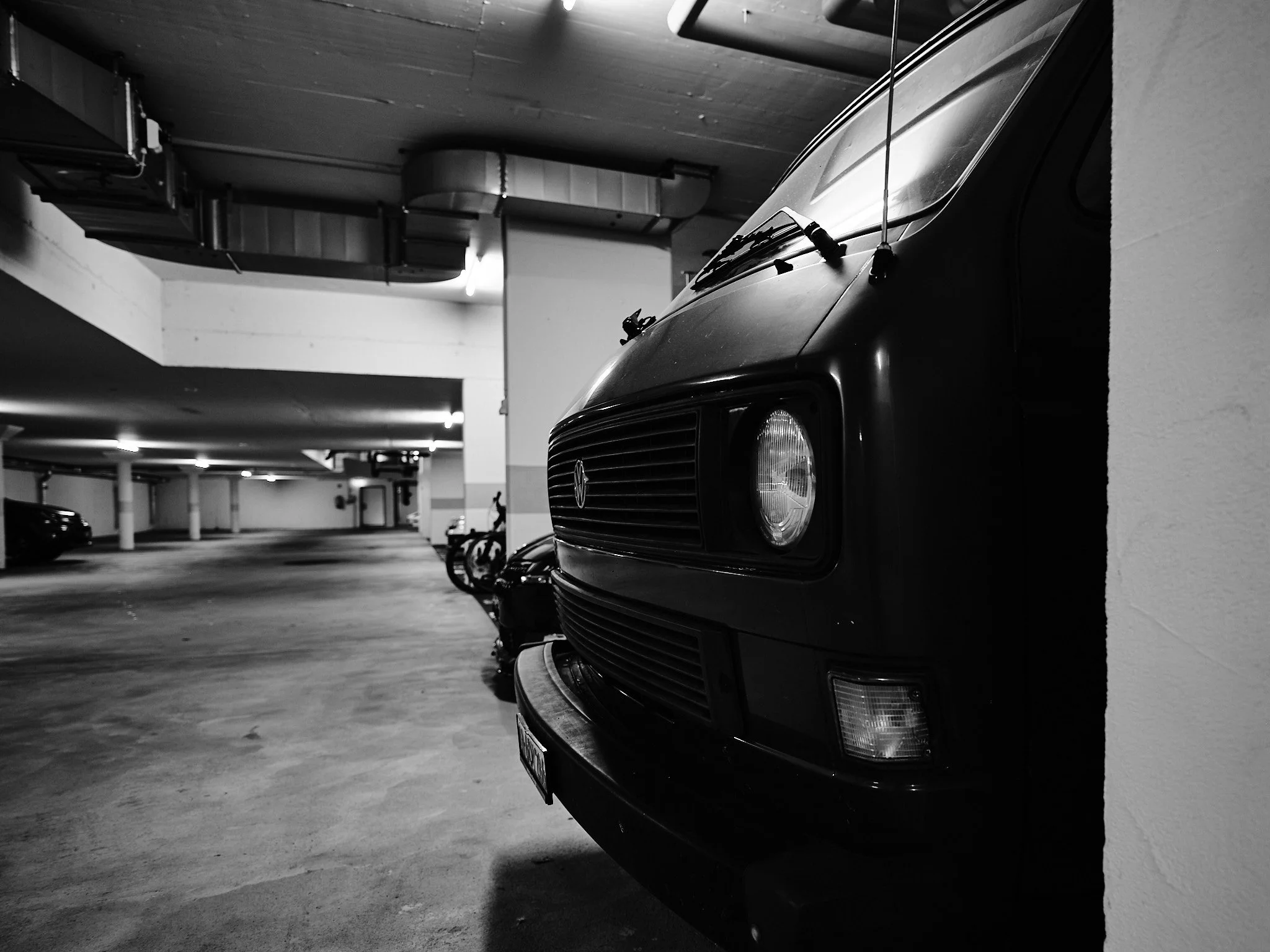 Black and white photo of a Volkswagen van parked in an indoor parking garage, with other cars and motorcycles in the background.