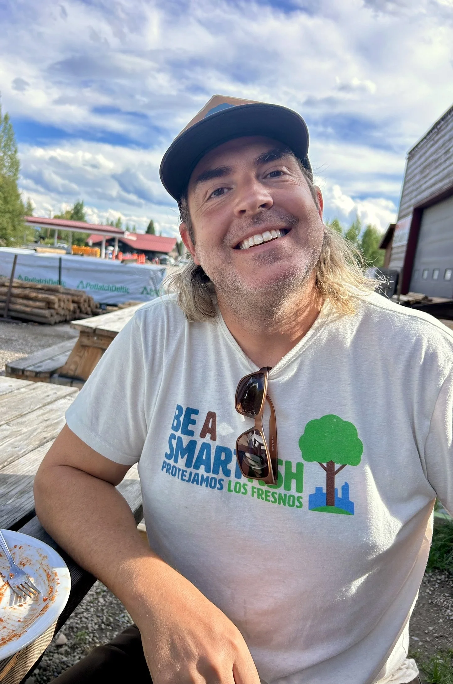 A man smiling and sitting outdoors at a wooden table, wearing a white t-shirt with a slogan and a graphic of trees and cityscape, sunglasses hanging from his shirt, under a partly cloudy sky.
