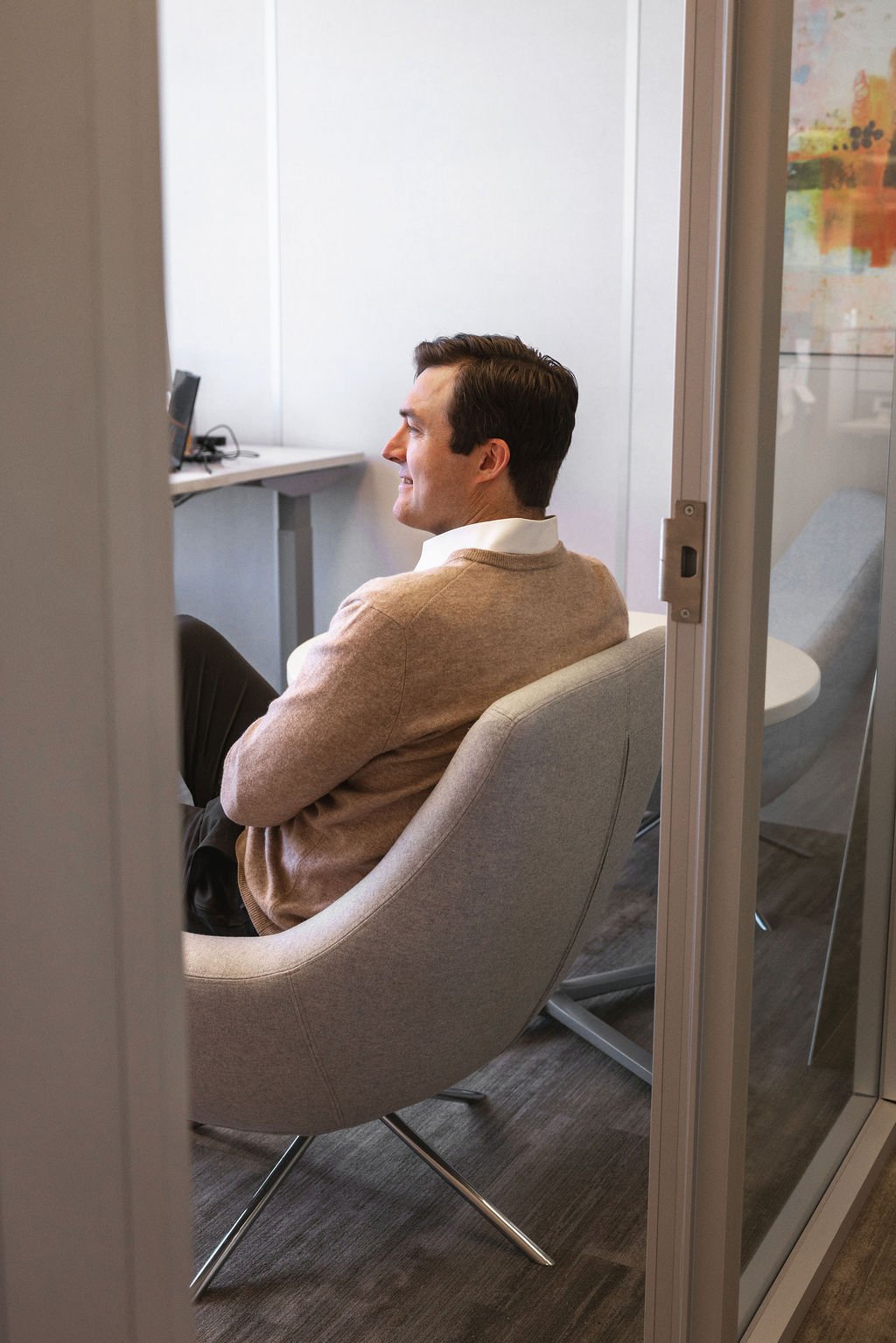 A man sitting in an office chair, smiling, viewed through a glass door. He has dark hair and is wearing a light brown sweater over a white shirt.