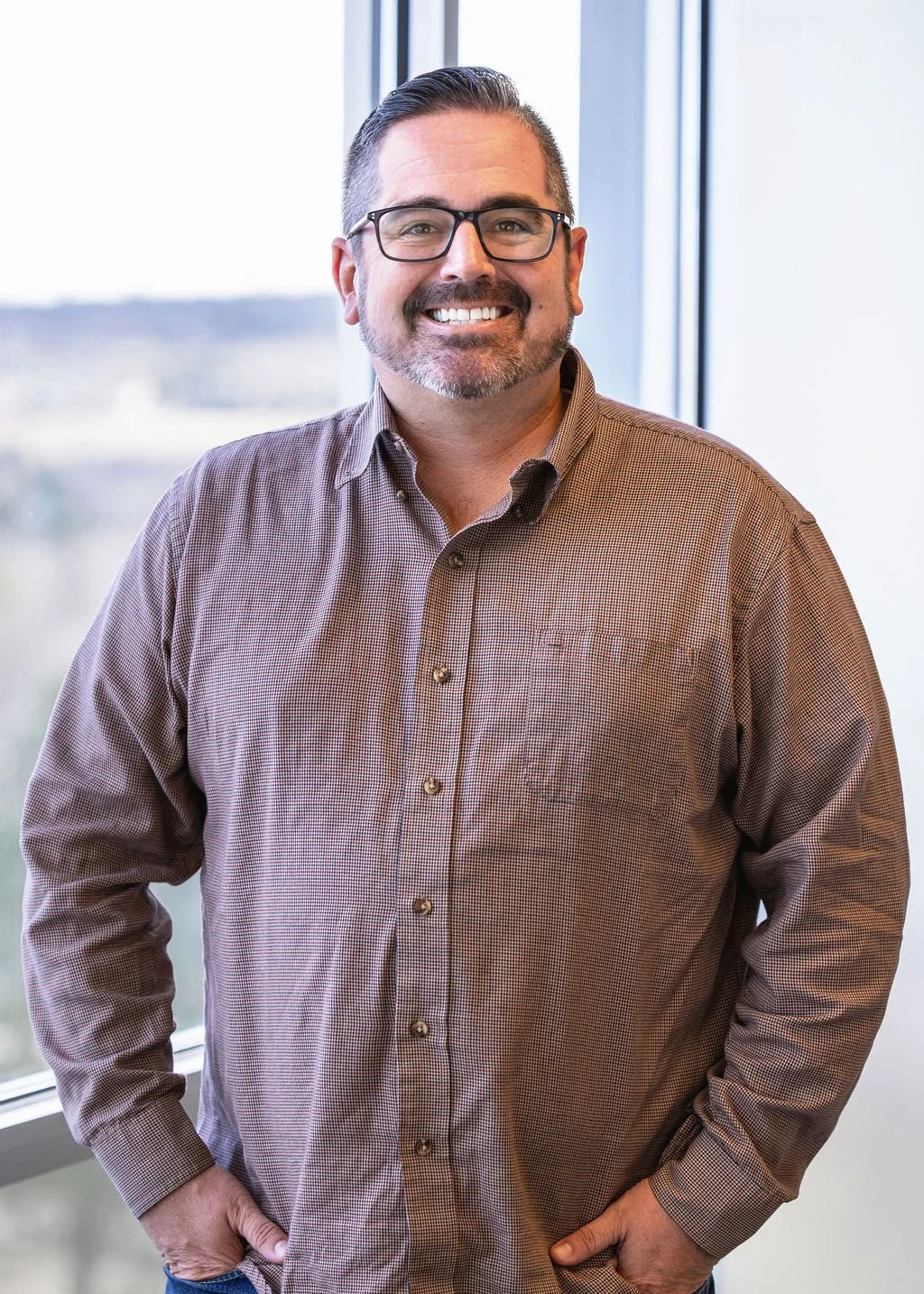 Man with glasses, beard, and short hair standing in front of a window, smiling, wearing a brown checkered button-up shirt.