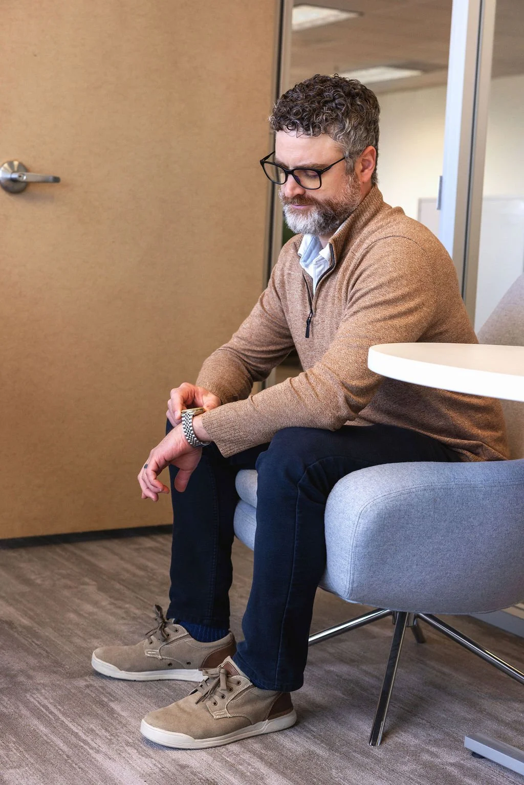 A middle-aged man with curly hair and glasses sits on a modern office chair, looking at his watch in a professional setting.