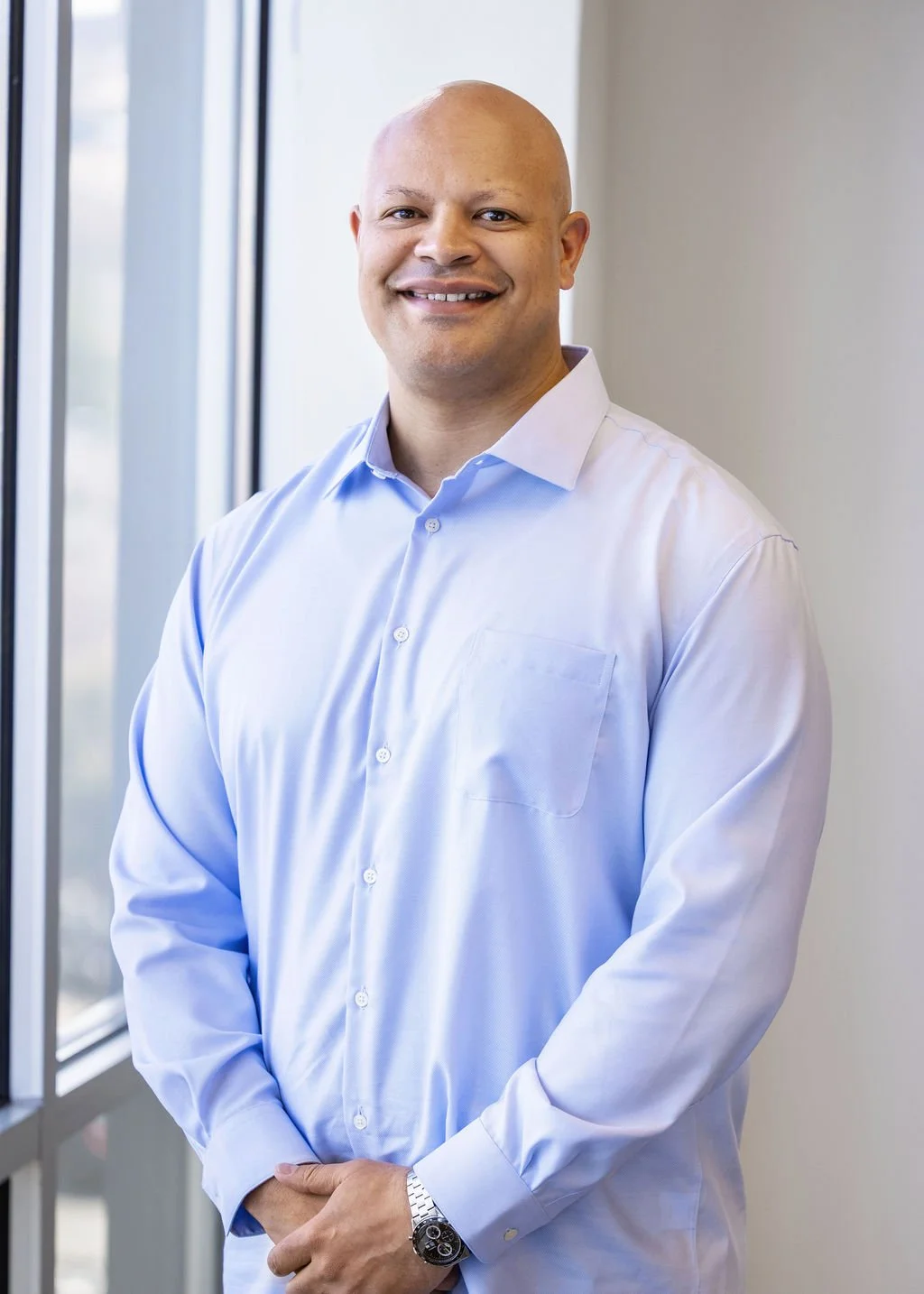 A smiling bald man wearing a light blue dress shirt, standing indoors near a window.