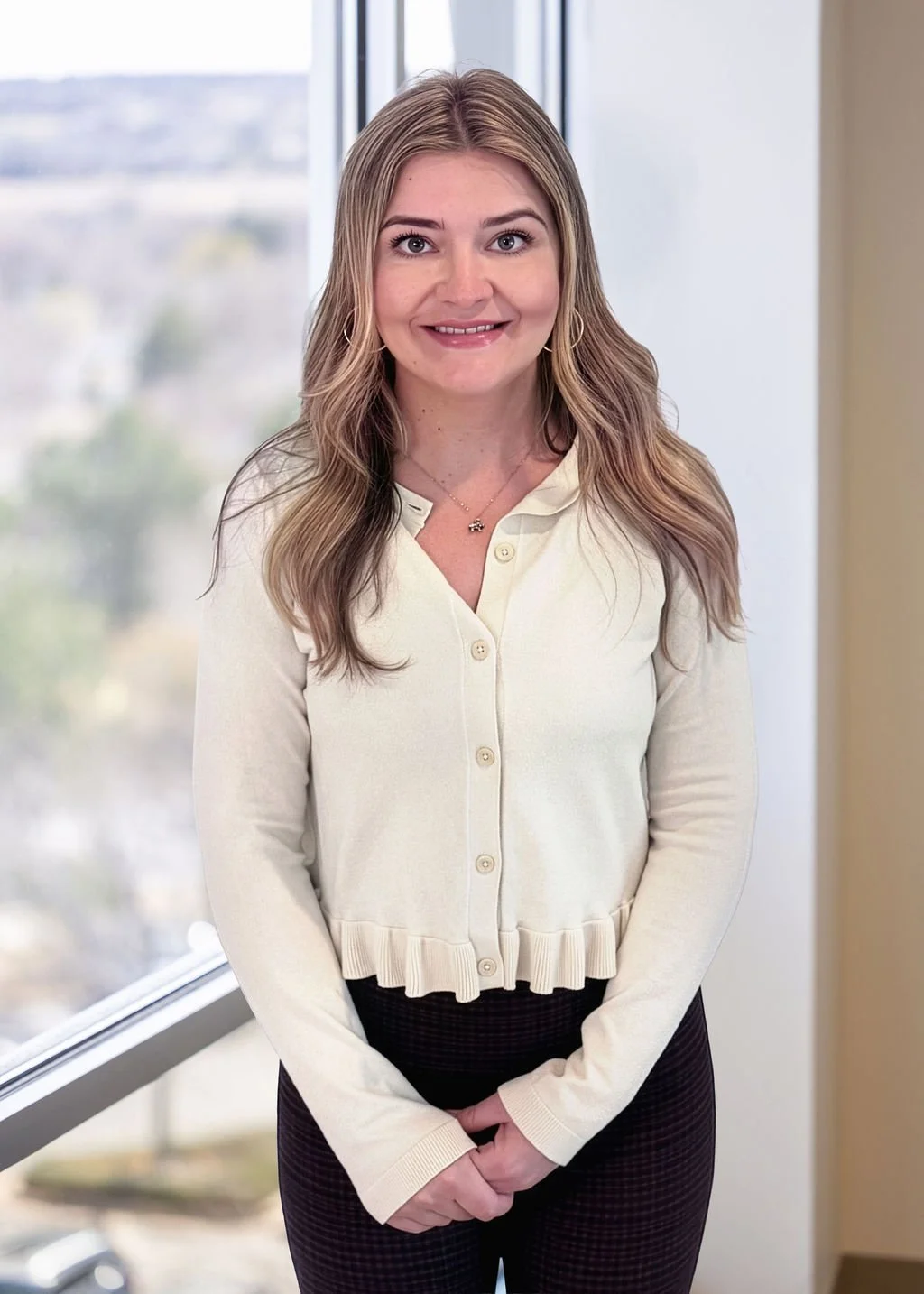 A young woman with long, wavy blonde hair smiling, standing indoors next to a large window with a cityscape in the background.