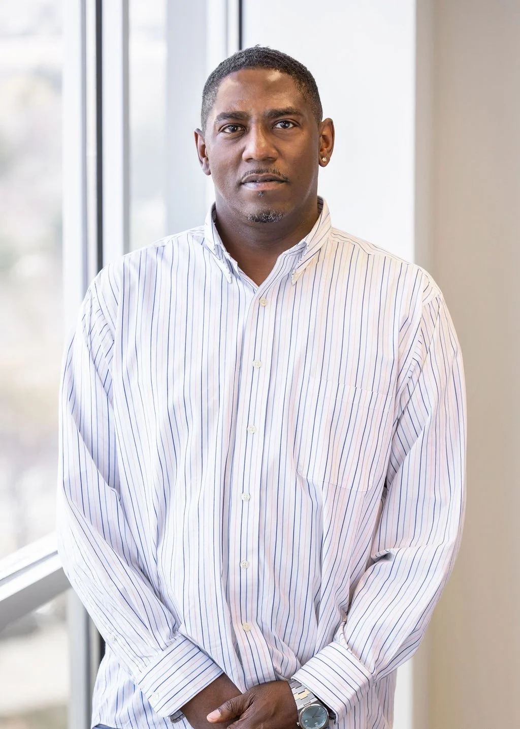 A man with short black hair and earrings wearing a white striped button-up shirt standing indoors near a window.