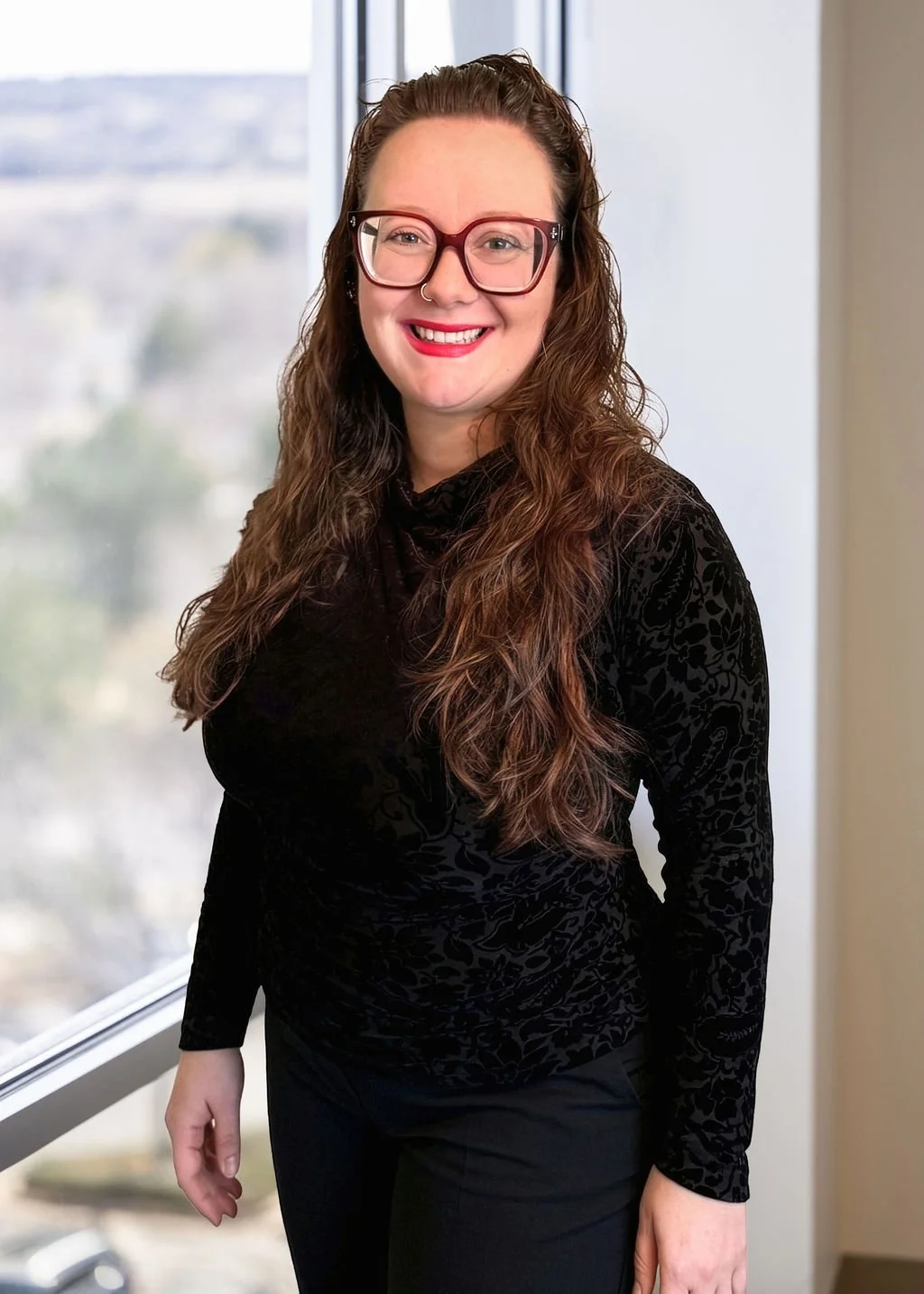 A woman with long curly brown hair, wearing glasses and a black textured top, standing by a window with a city view outside, smiling at the camera.