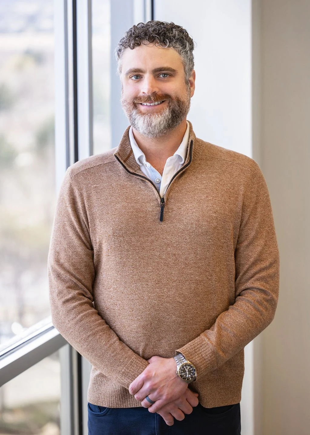 A man standing indoors near a window, smiling at the camera, wearing a tan sweater over a white shirt, with curly hair and a beard.