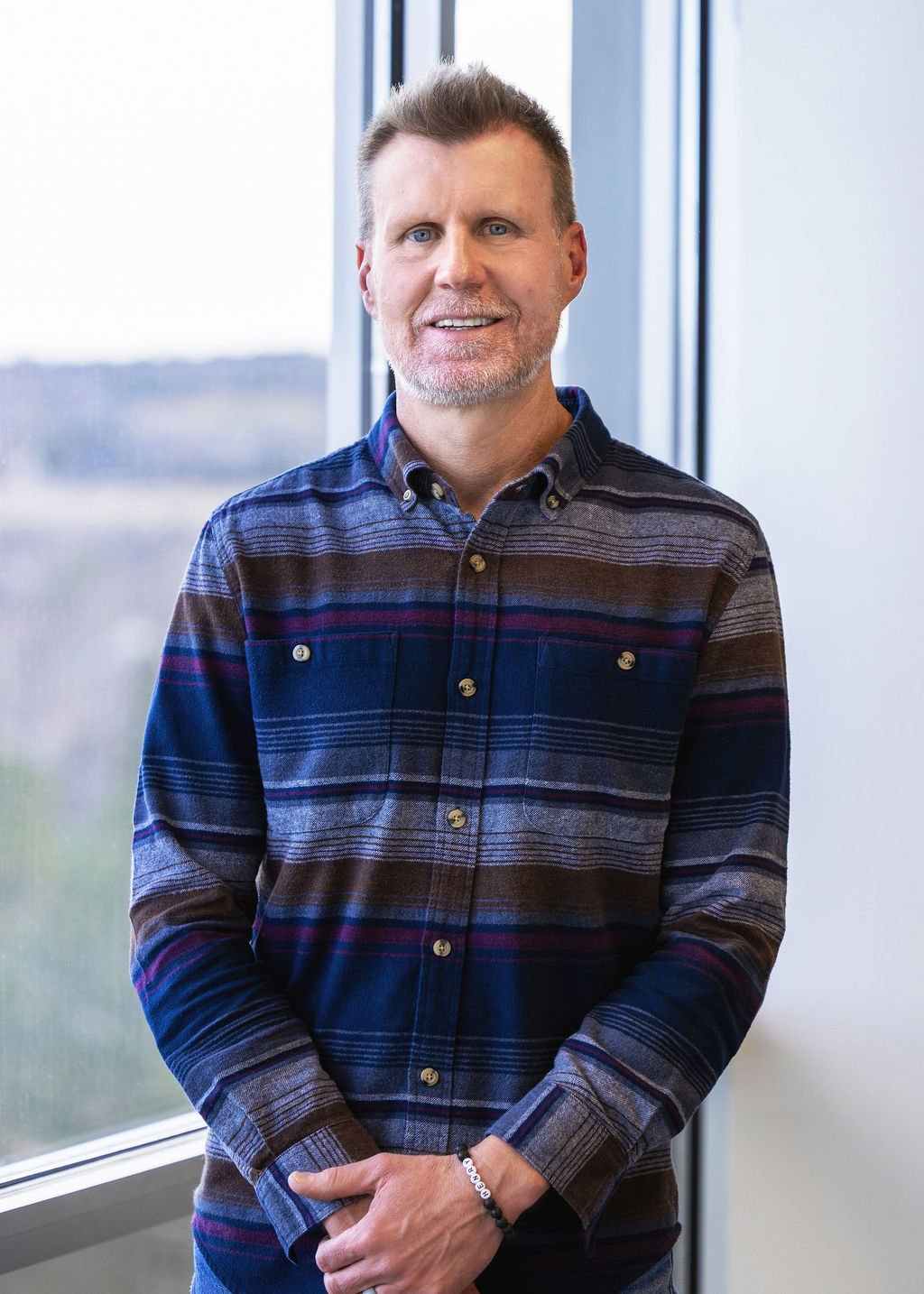 A smiling man with short gray hair and a beard, wearing a dark plaid shirt, standing indoors near a large window with a blurred outdoor background.
