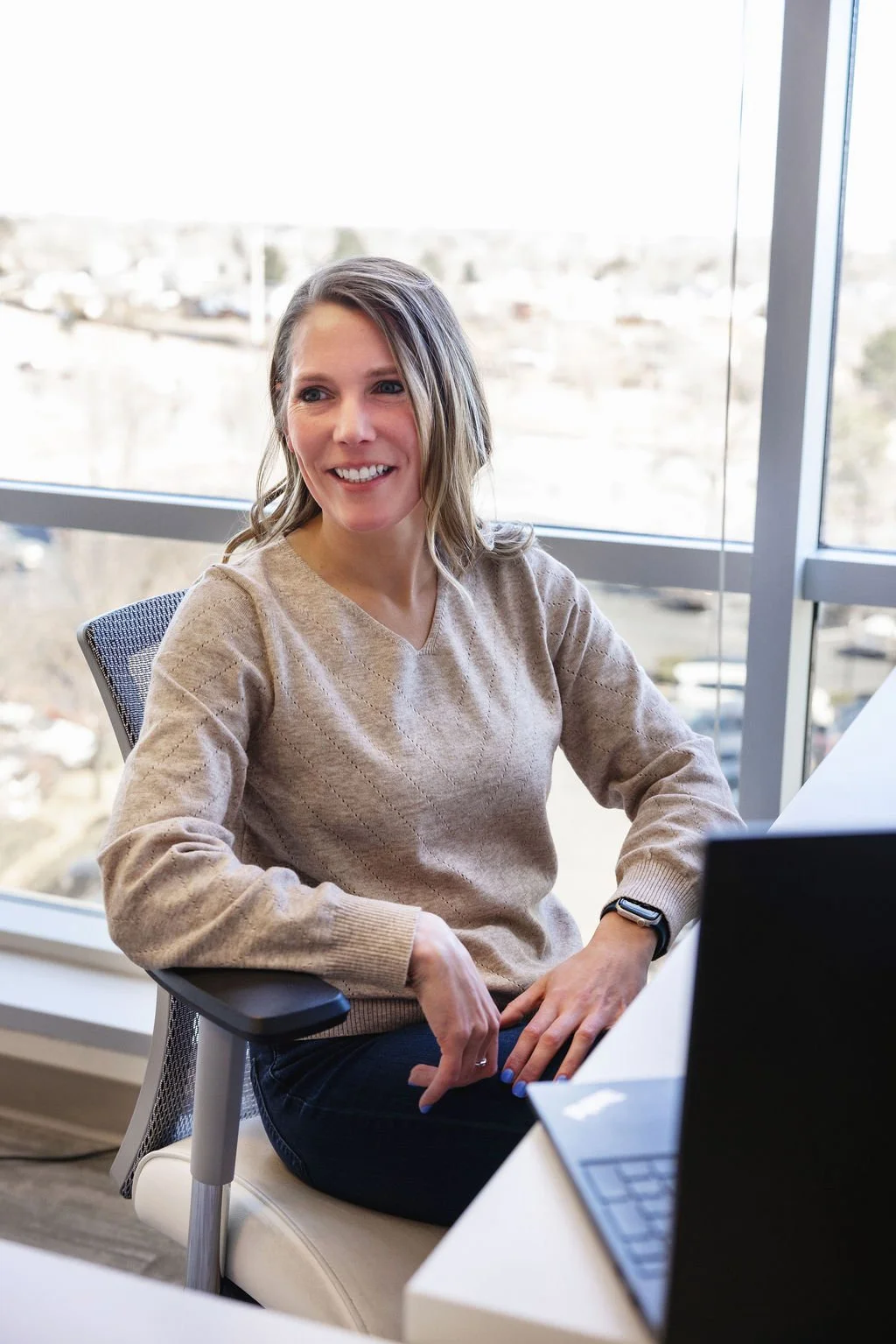 Woman with shoulder-length hair smiling while sitting at an office desk near large windows with a city view.