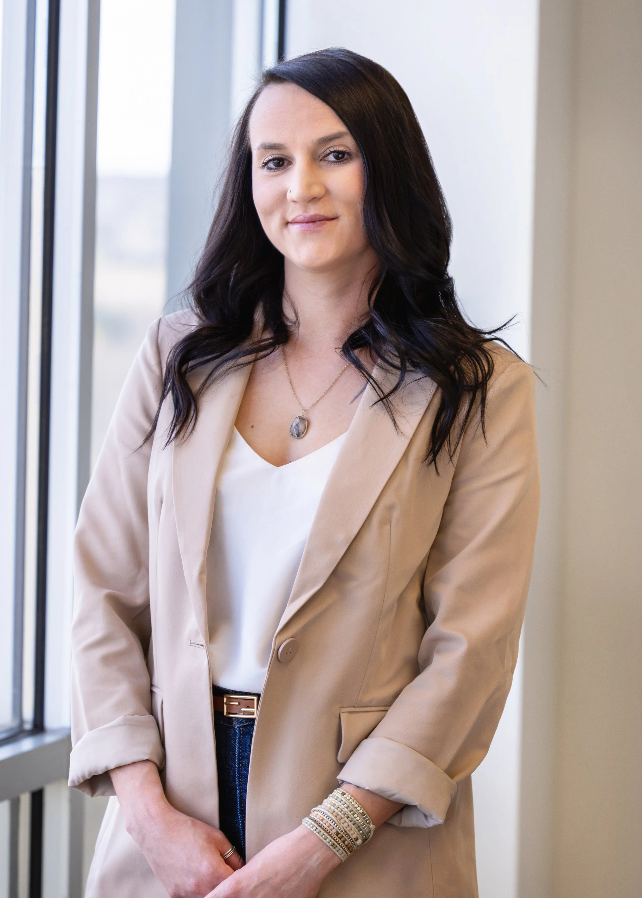 A woman with long dark wavy hair, wearing a light beige blazer over a white top, standing near a window in a modern office.