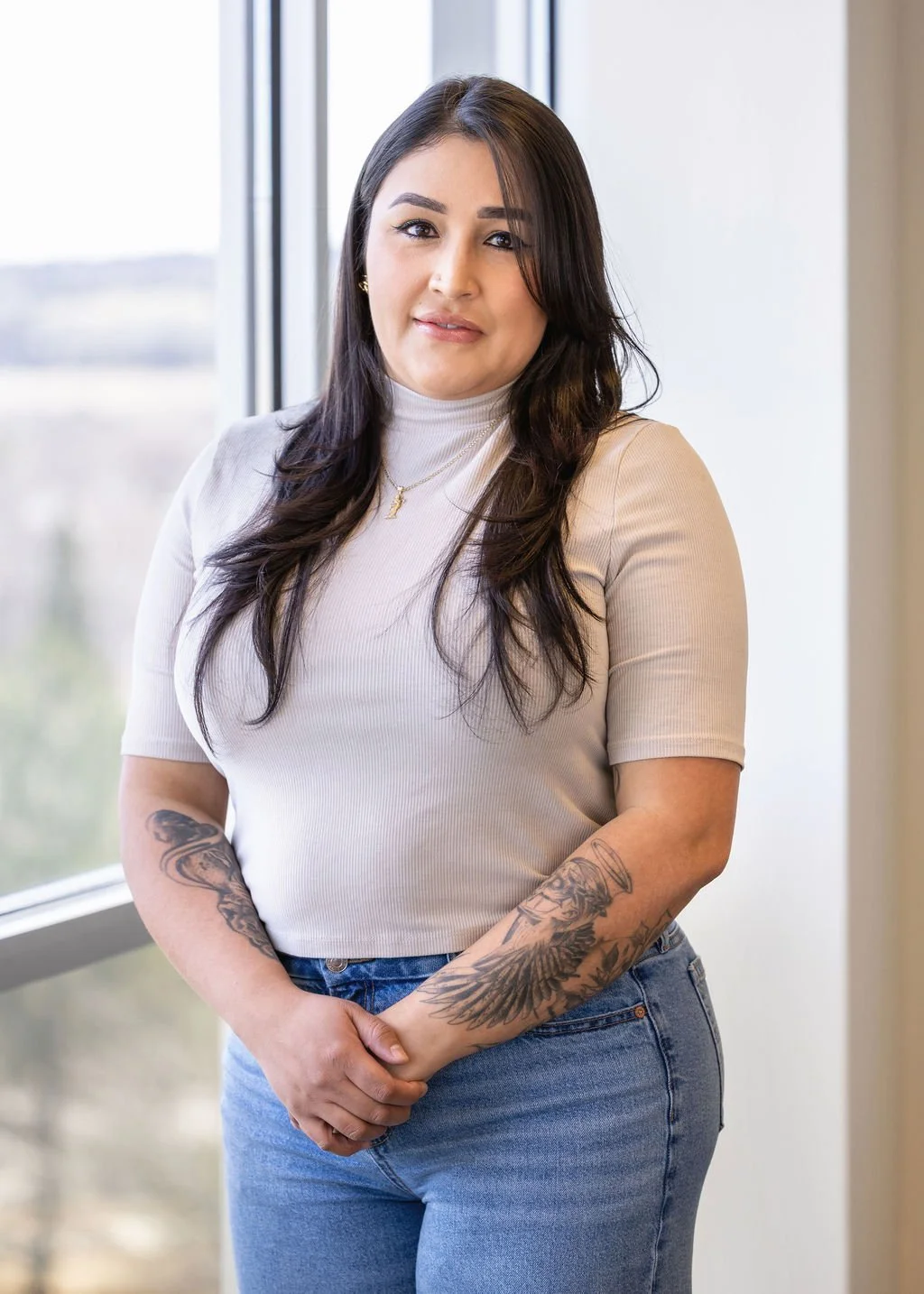 A woman with long dark hair, tattoos on her arms, wearing a white turtleneck and blue jeans, standing indoors near a window.