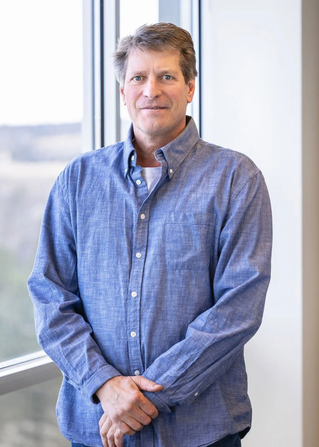 A middle-aged man with light brown hair, blue eyes, and fair skin, standing indoors with a large window behind him. He is wearing a blue button-down shirt over a white undershirt and is looking at the camera with a slight smile.