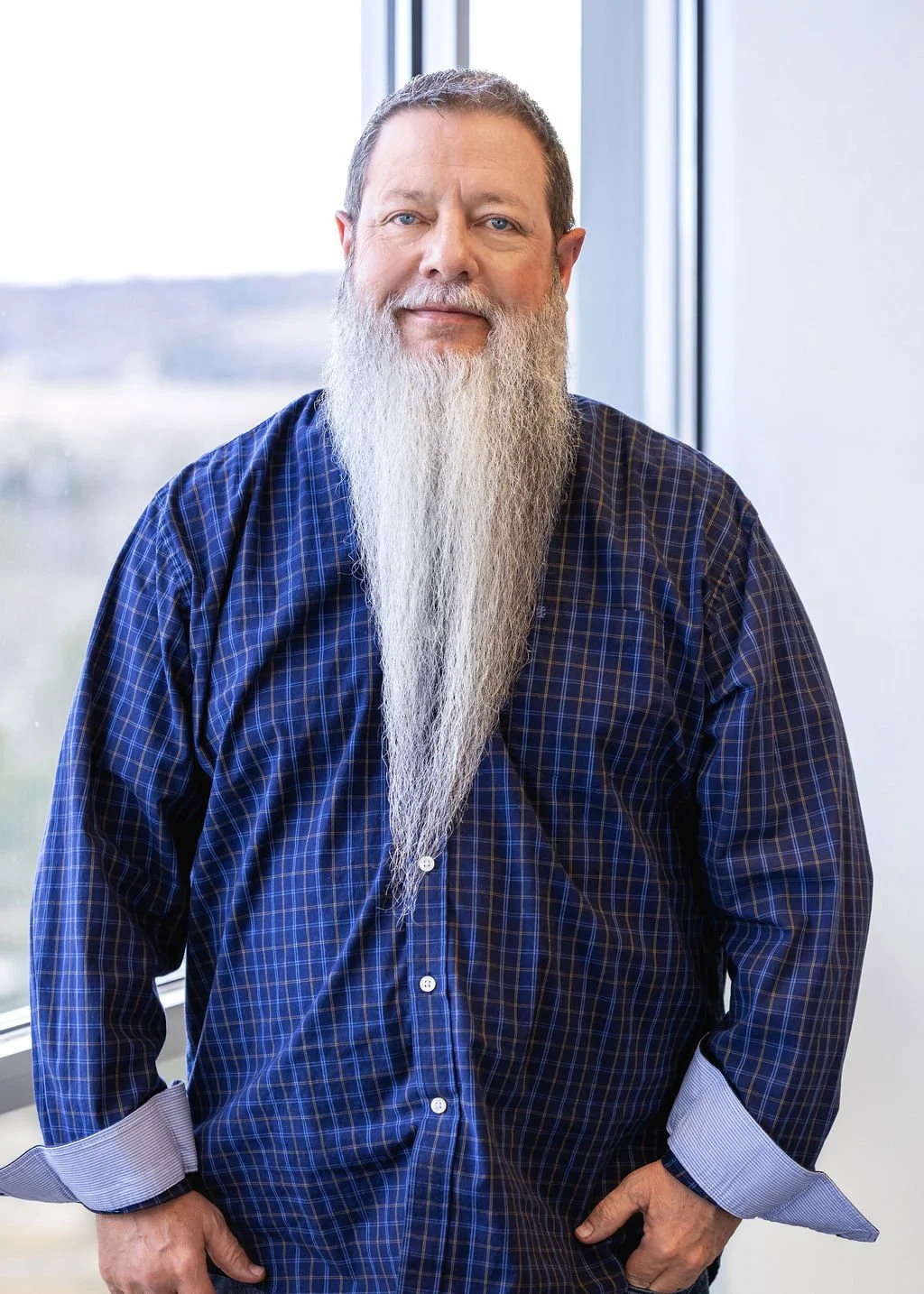 A man with a long white beard wearing a blue checkered shirt, standing indoors near a large window.