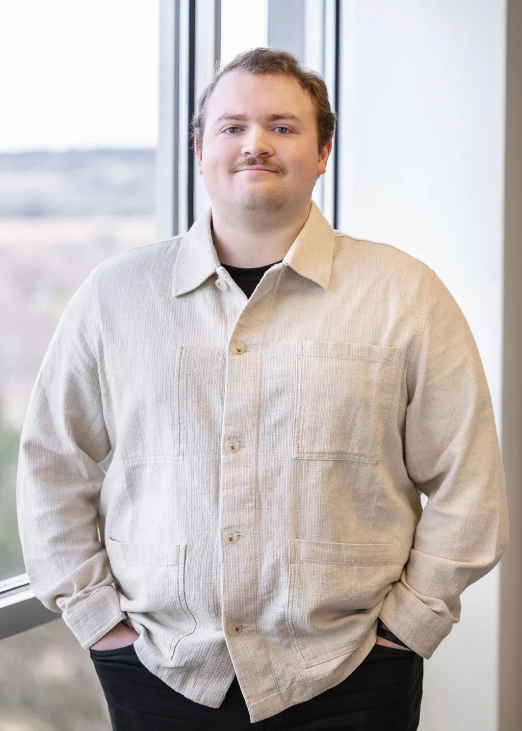 A man standing indoors near a large window, wearing a beige shirt and black pants, with his hands in his pockets, smiling at the camera.