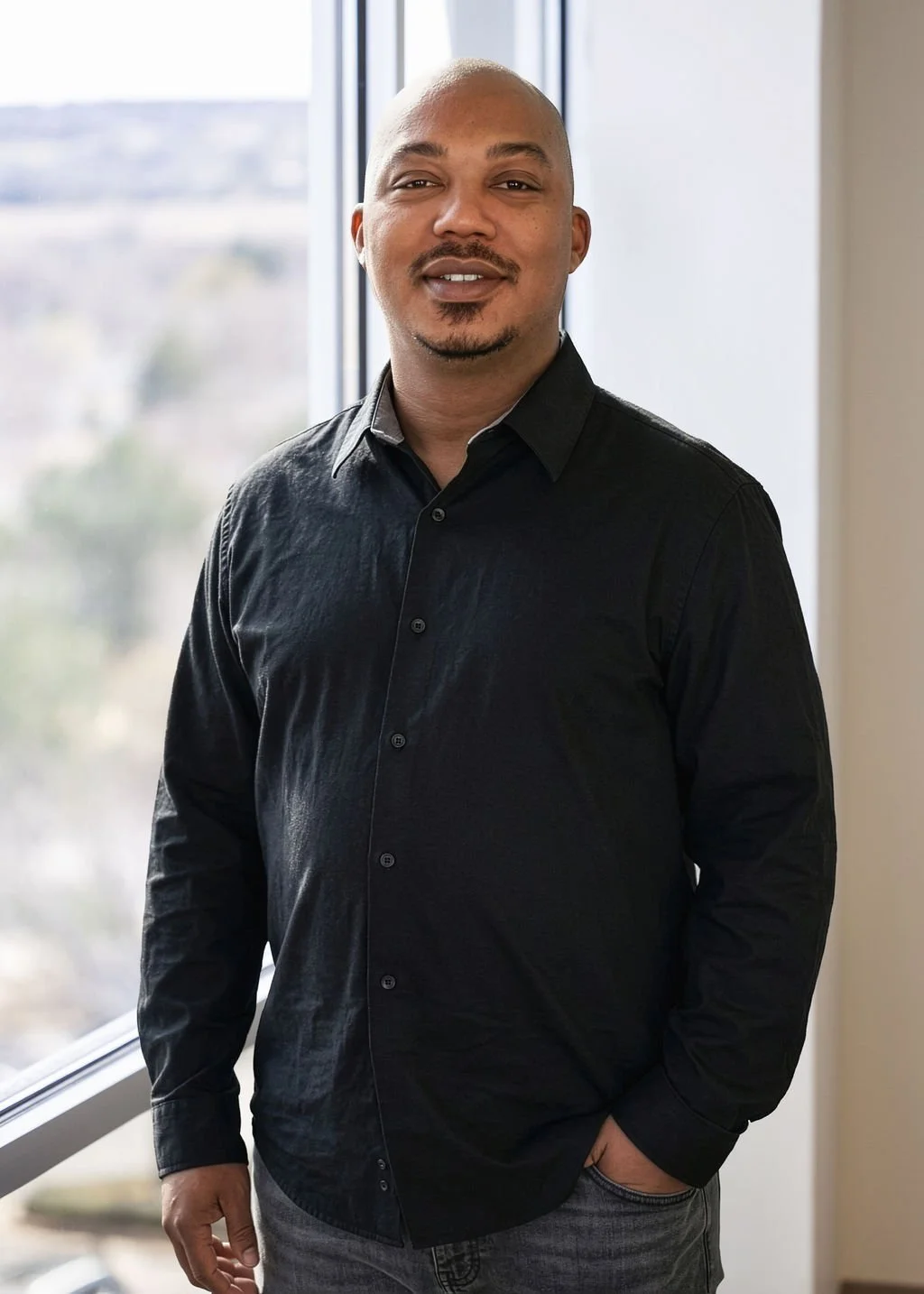 A man with a shaved head and a goatee, wearing a black button-up shirt, standing indoors by a large window with a cityscape in the background.