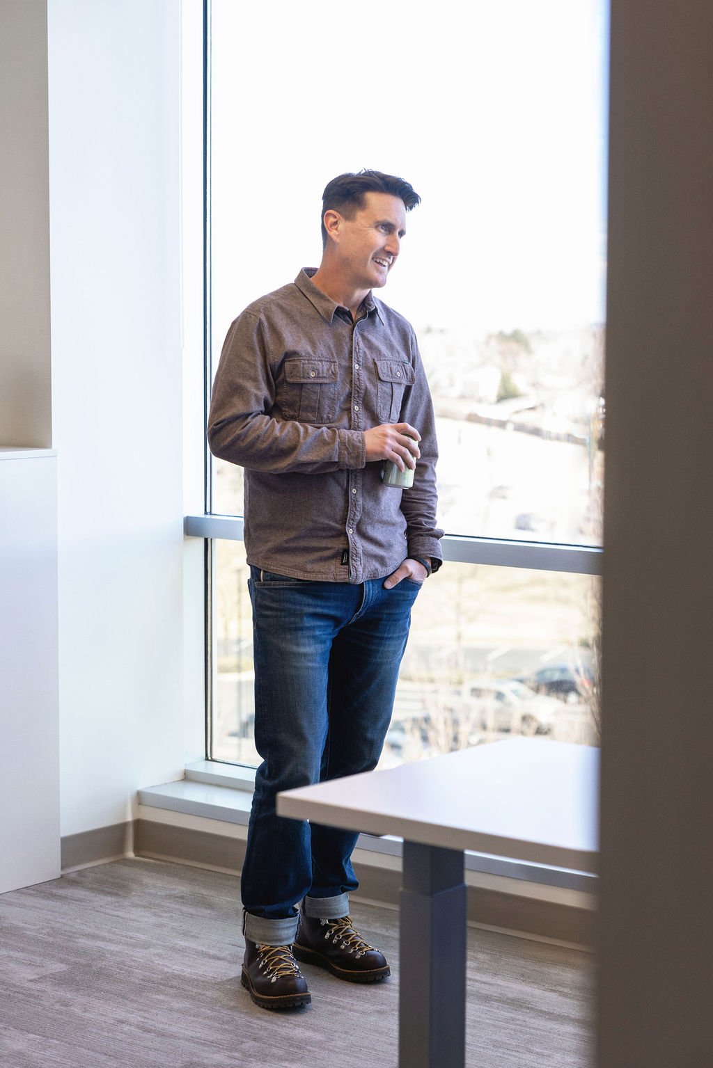 A young man standing by a large window, smiling and holding a drink, wearing a brown shirt, jeans, and boots, in an office setting.
