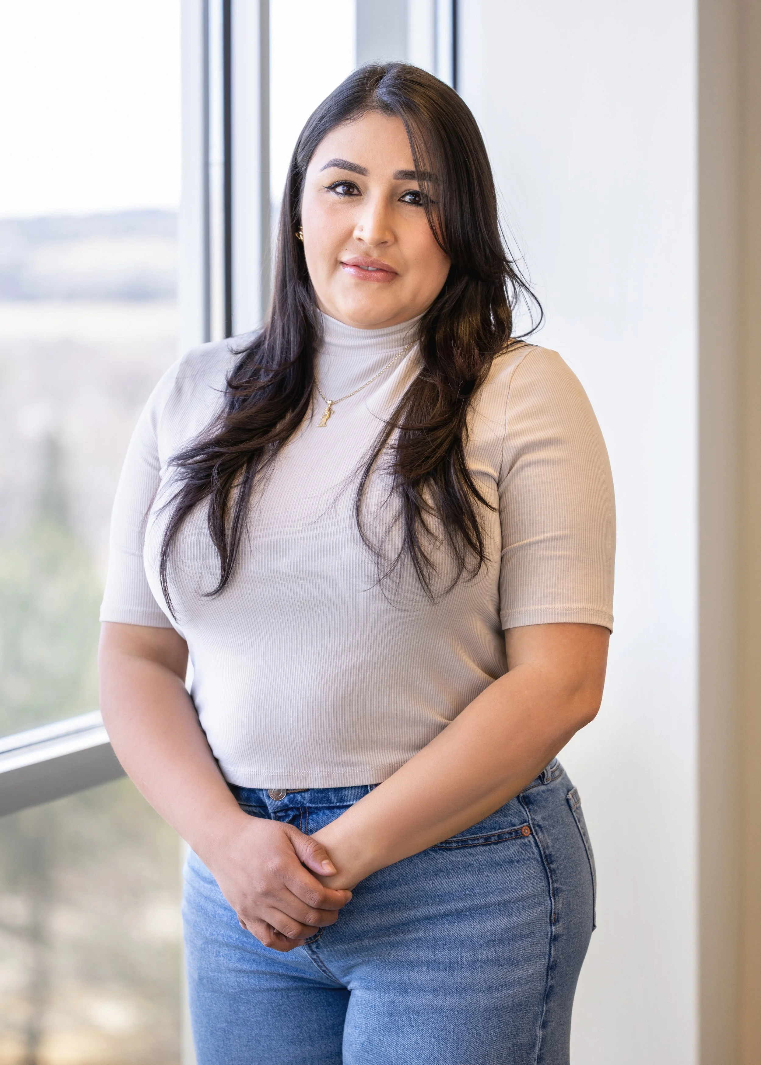 A young woman with long dark hair, wearing a white turtleneck shirt and blue jeans, standing near a window with her hands clasped in front. She has tattoos on her arms and is looking at the camera with a slight smile.