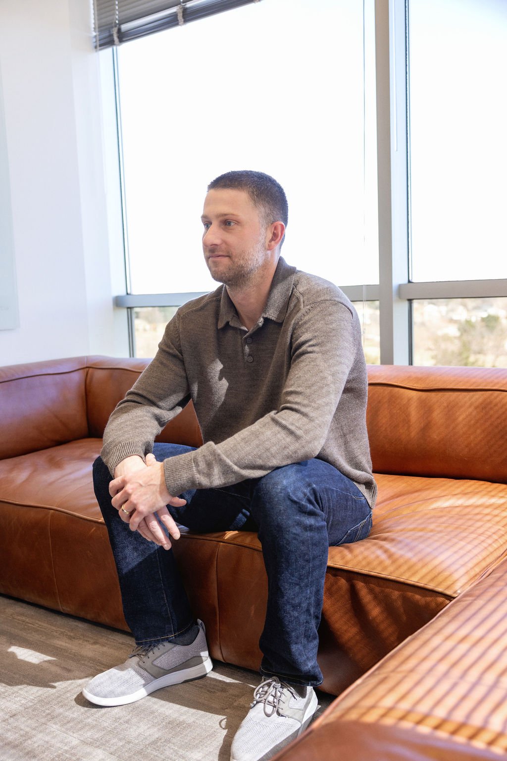 A man with short dark hair and a beard sitting on a tan leather sofa in front of large windows, dressed in a brown sweater, dark jeans, and gray sneakers.