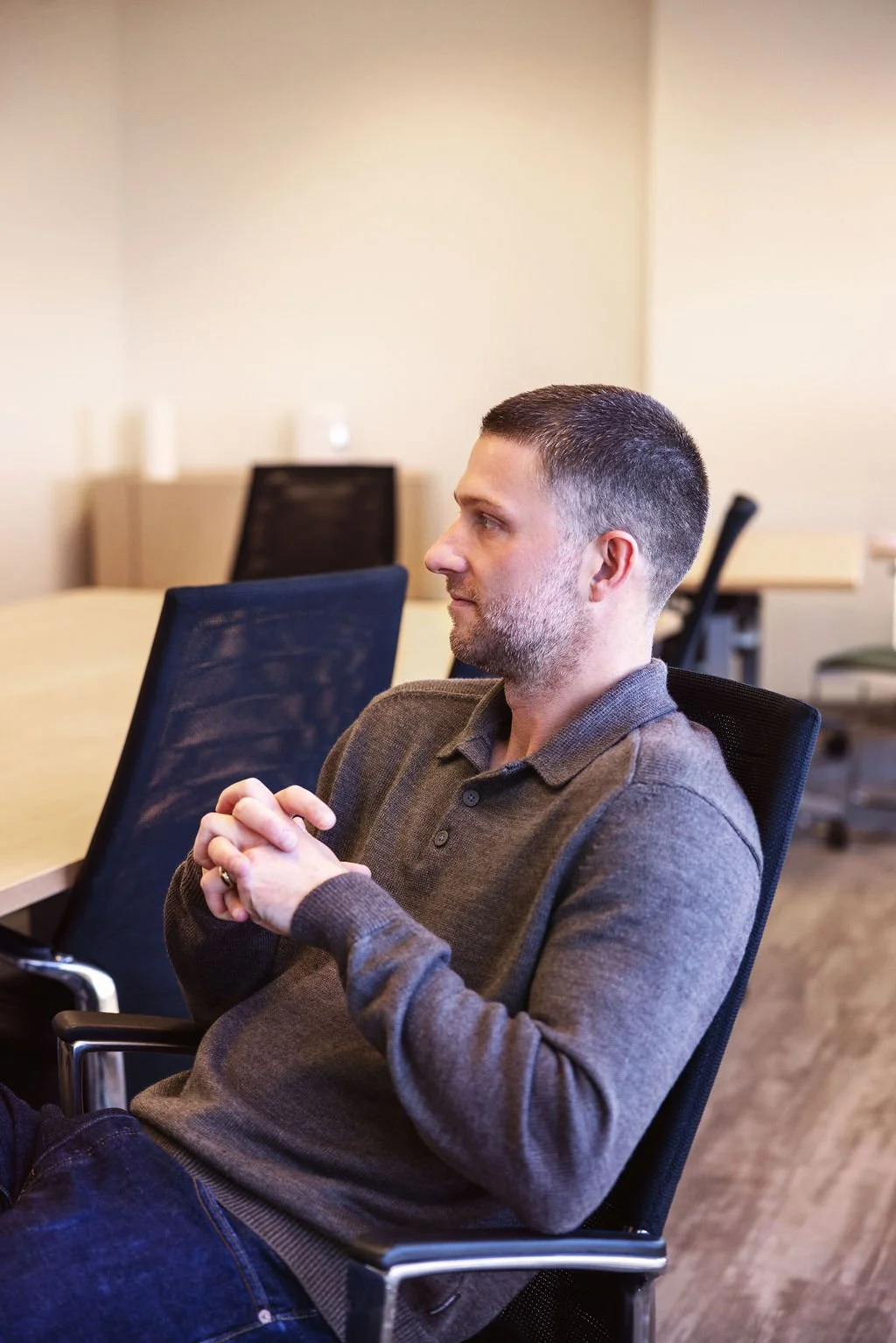 A man with short dark hair, a beard, wearing a brown sweater, sitting in a black office chair in a meeting room.