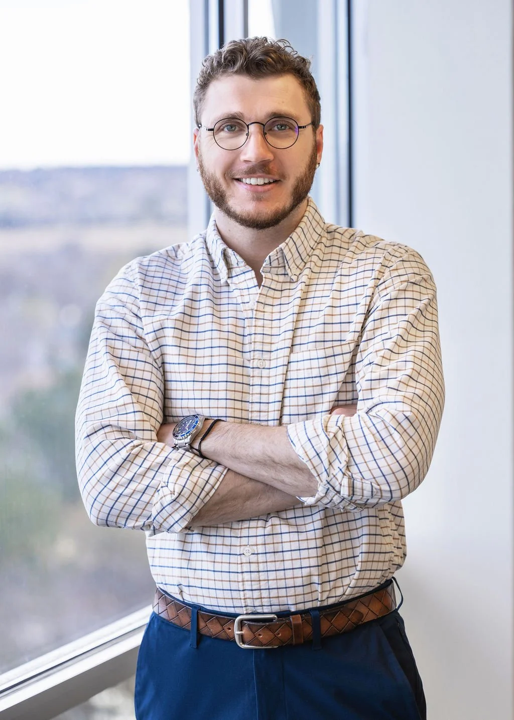 A young man with glasses, a beard, and curly hair, standing in front of a large window with cityscape view, smiling, wearing a checkered dress shirt, a watch, and a leather belt.