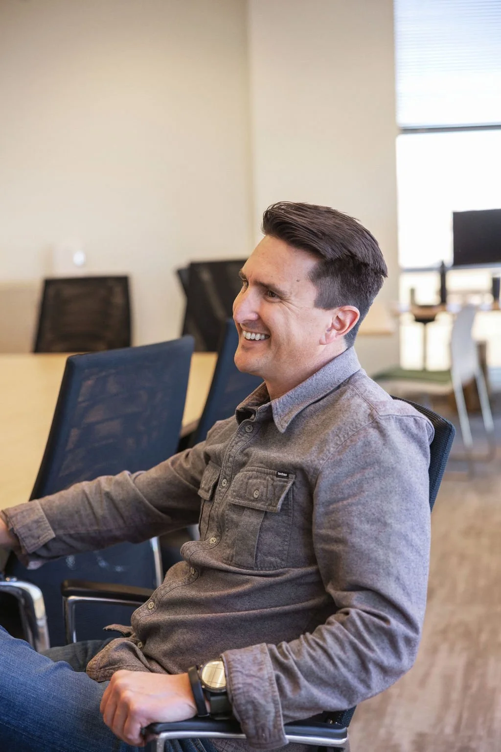 A young man in a gray shirt sitting in a conference room, smiling, with chairs and a table in the background.