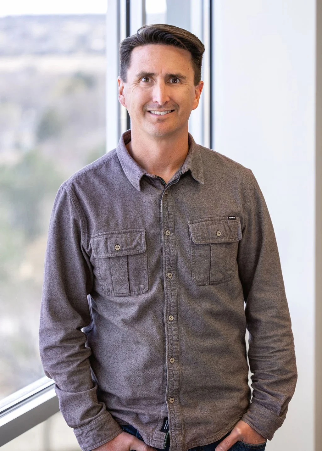 A man standing indoors near a window, smiling at the camera, wearing a gray button-up shirt.