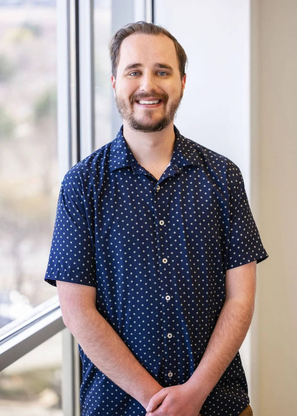 A smiling man with a beard and short hair, wearing a dark blue shirt with white polka dots, stands indoors near a window with a blurred outdoor background.