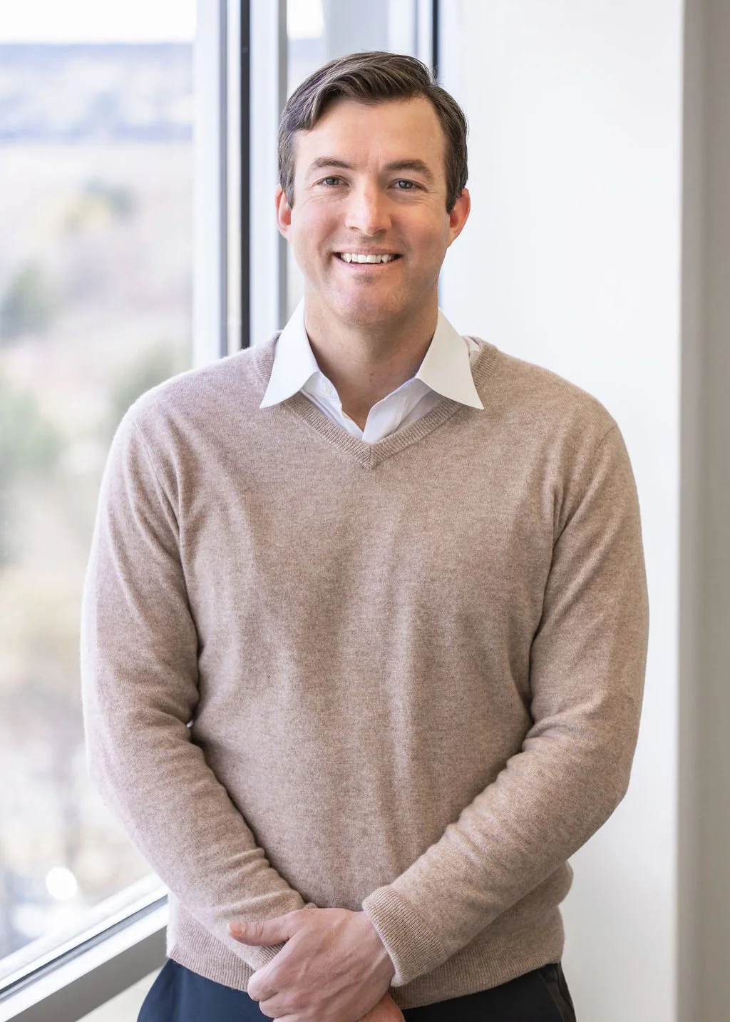 A smiling man standing indoors near a window, wearing a white shirt and beige sweater.