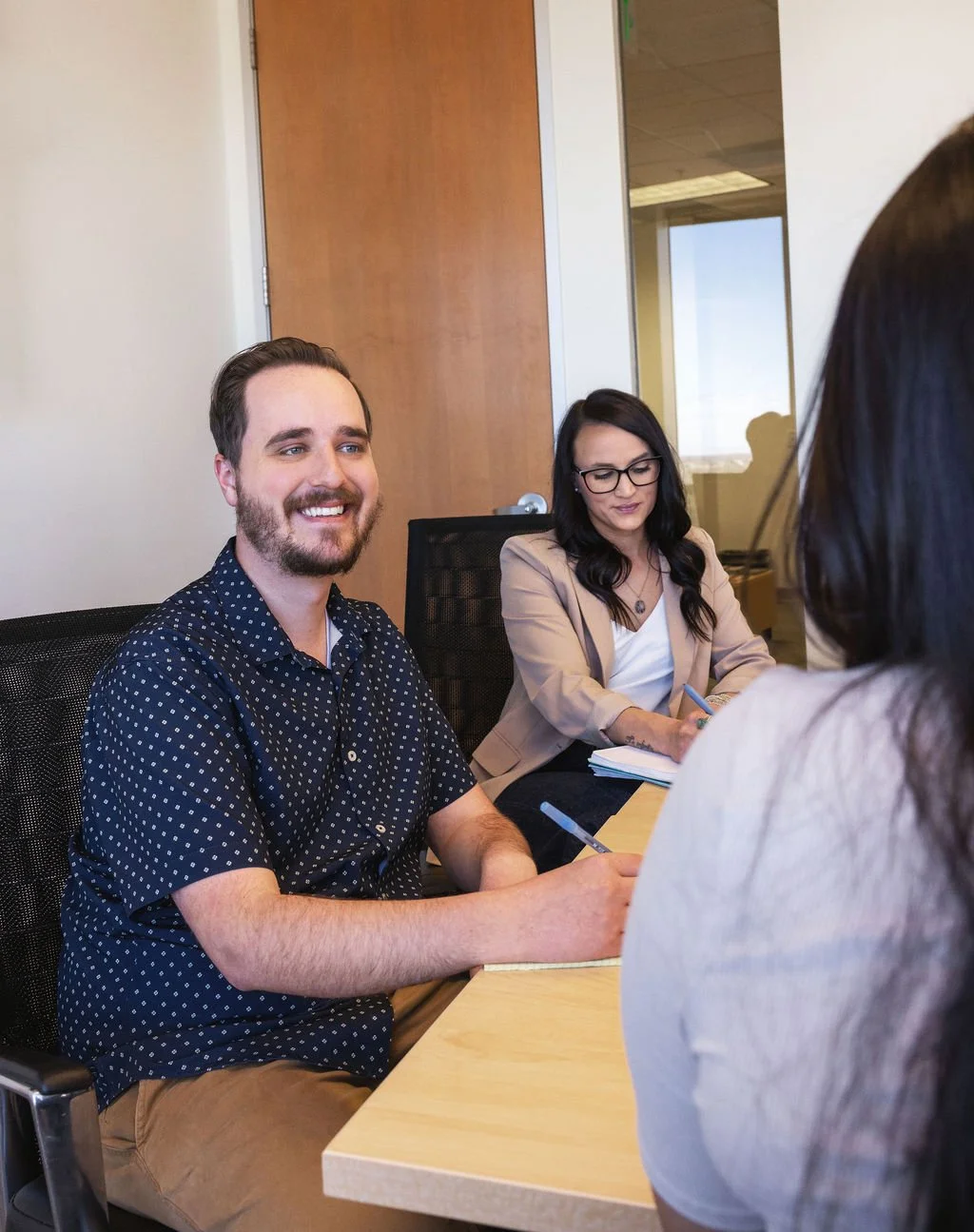 A group of four people in a meeting room, with one woman taking notes, two women partially visible, and a man smiling and looking ahead.