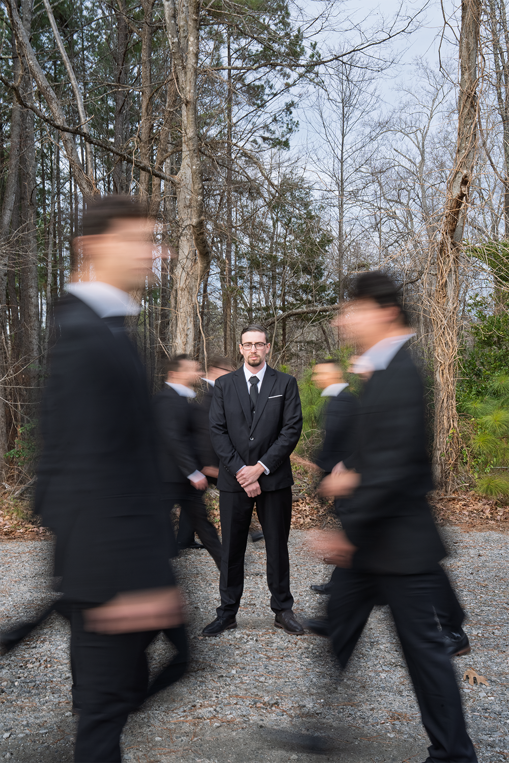 A man in a black suit and glasses stands still amidst a group of other men in suits walking past him on a gravel path in a wooded area with bare trees.