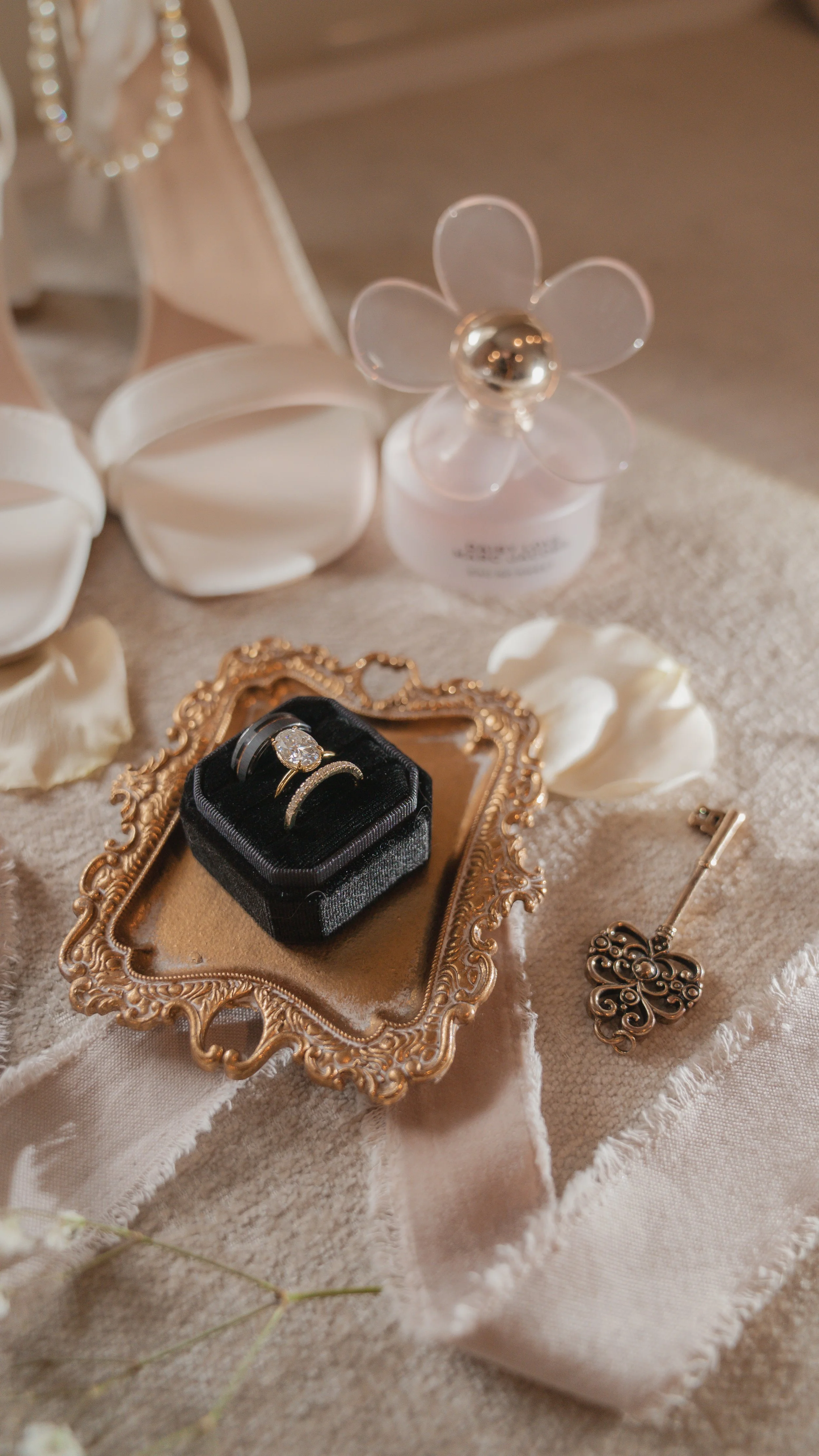 A jewelry display with rings, a gold butterfly-shaped jewelry piece, and a perfume bottle, along with earrings and a white satin cloth on a beige surface, surrounded by white petals and a flower-shaped perfume bottle.