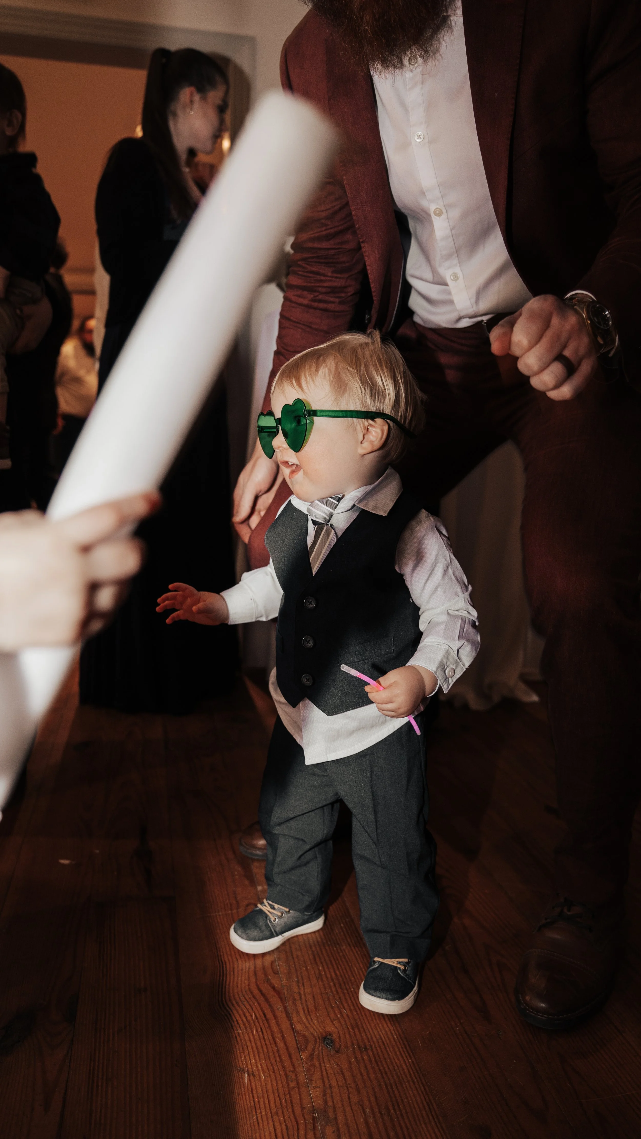 A young boy wearing green heart-shaped sunglasses, a gray vest, white shirt, and dark pants is dancing at a party. An adult man with a beard in a red suit and white shirt is clapping nearby. Other people are visible in the background.