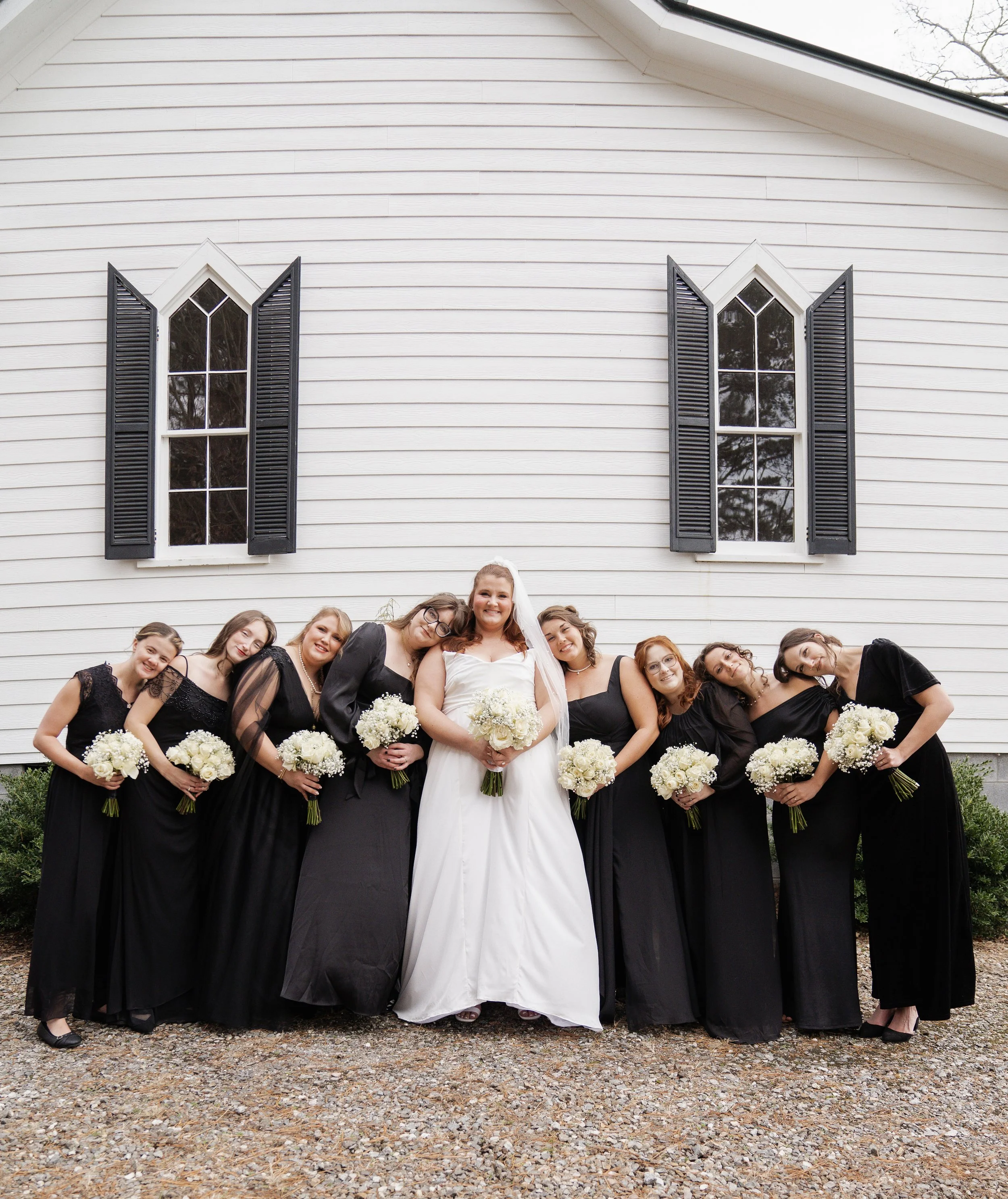 A bride in a white wedding dress standing with several bridesmaids dressed in black outside a white house with black shutters, all holding bouquets of white flowers.