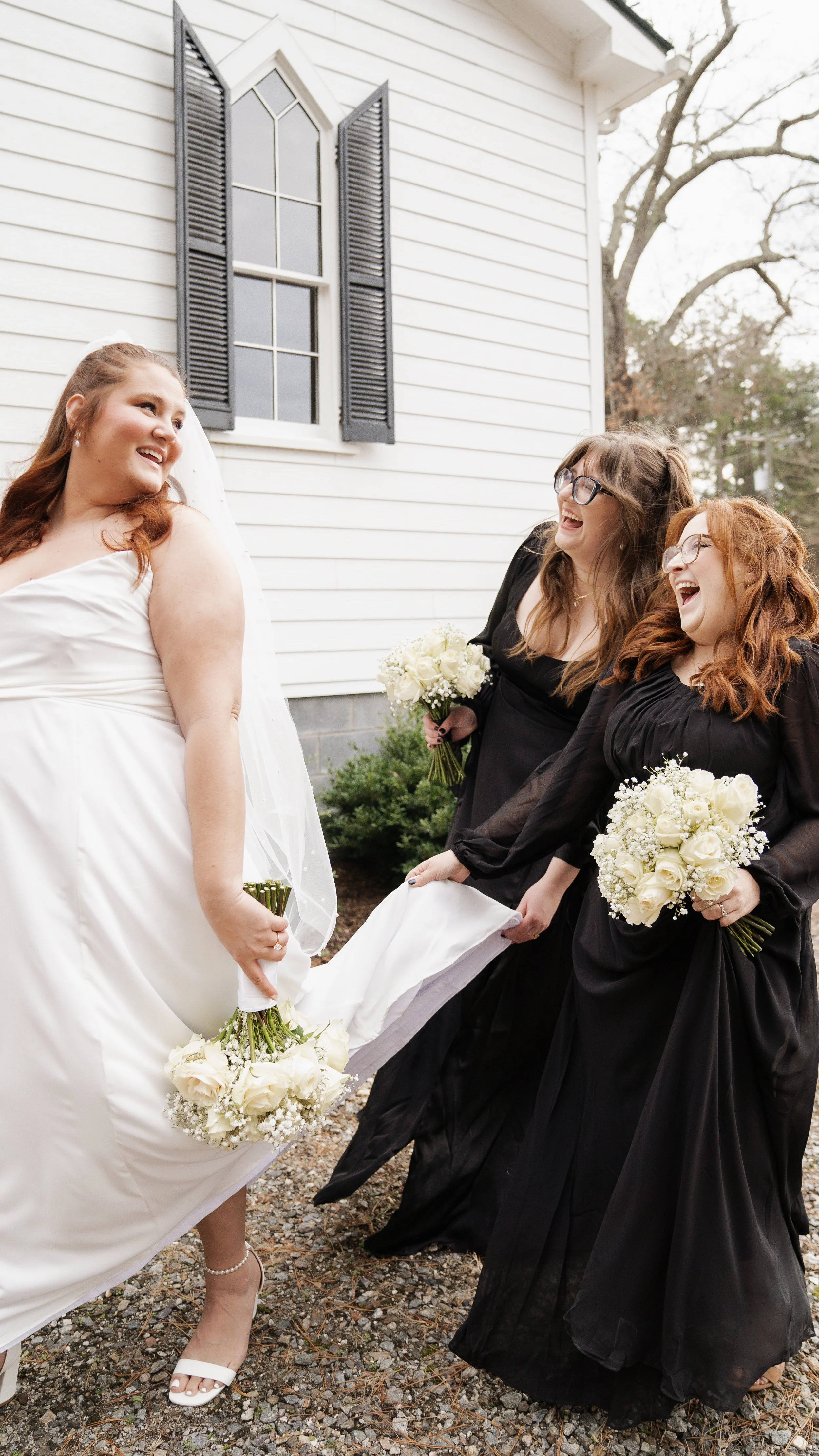 Three women, one bride in a white wedding dress and veil, and two bridesmaids in black dresses, are holding bouquets of white roses and smiling joyous outside a white house with black shutters.