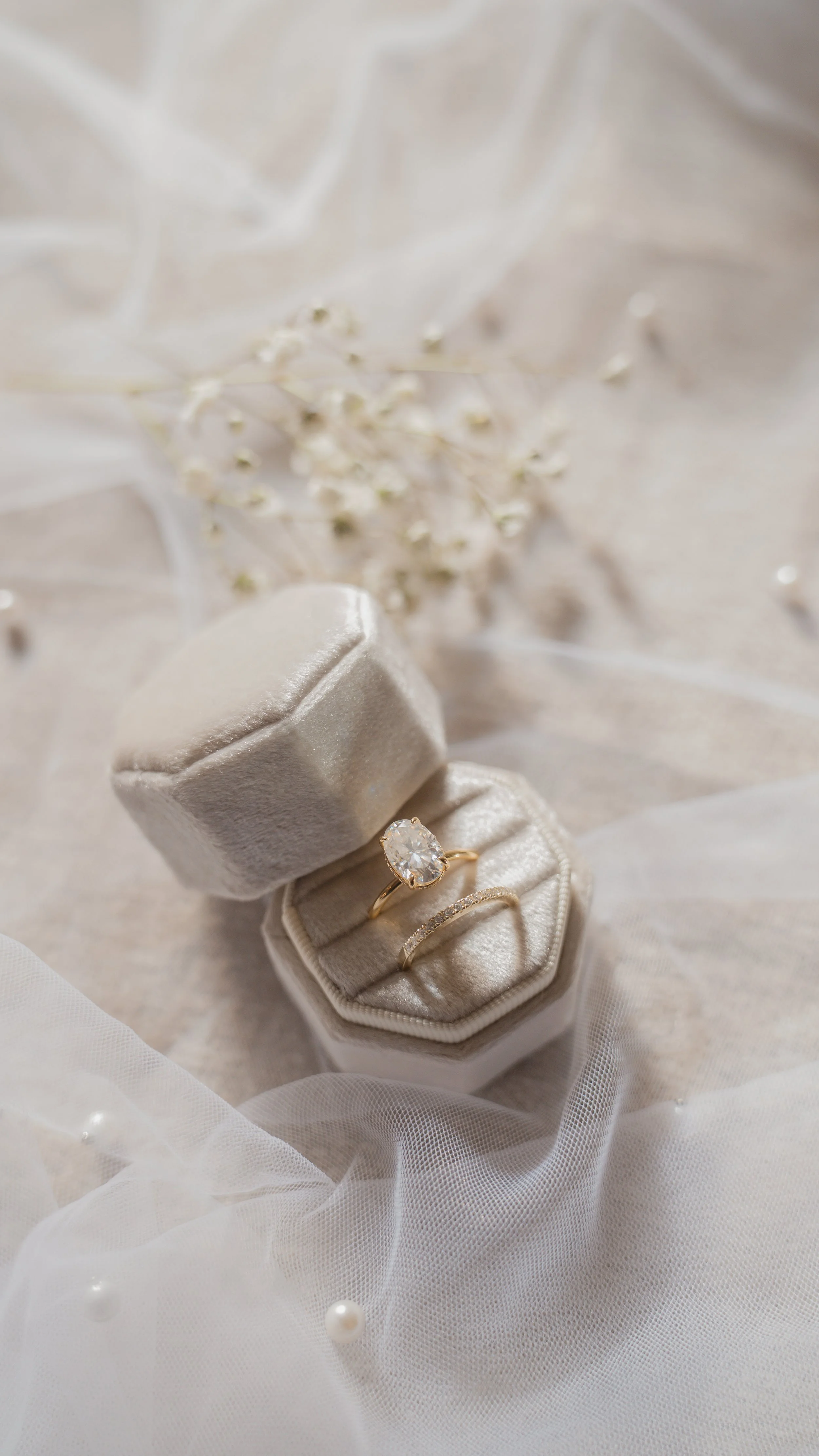 A gold ring with a large diamond in a velvet jewelry box surrounded by delicate white flowers and pearls, set on soft white fabric.