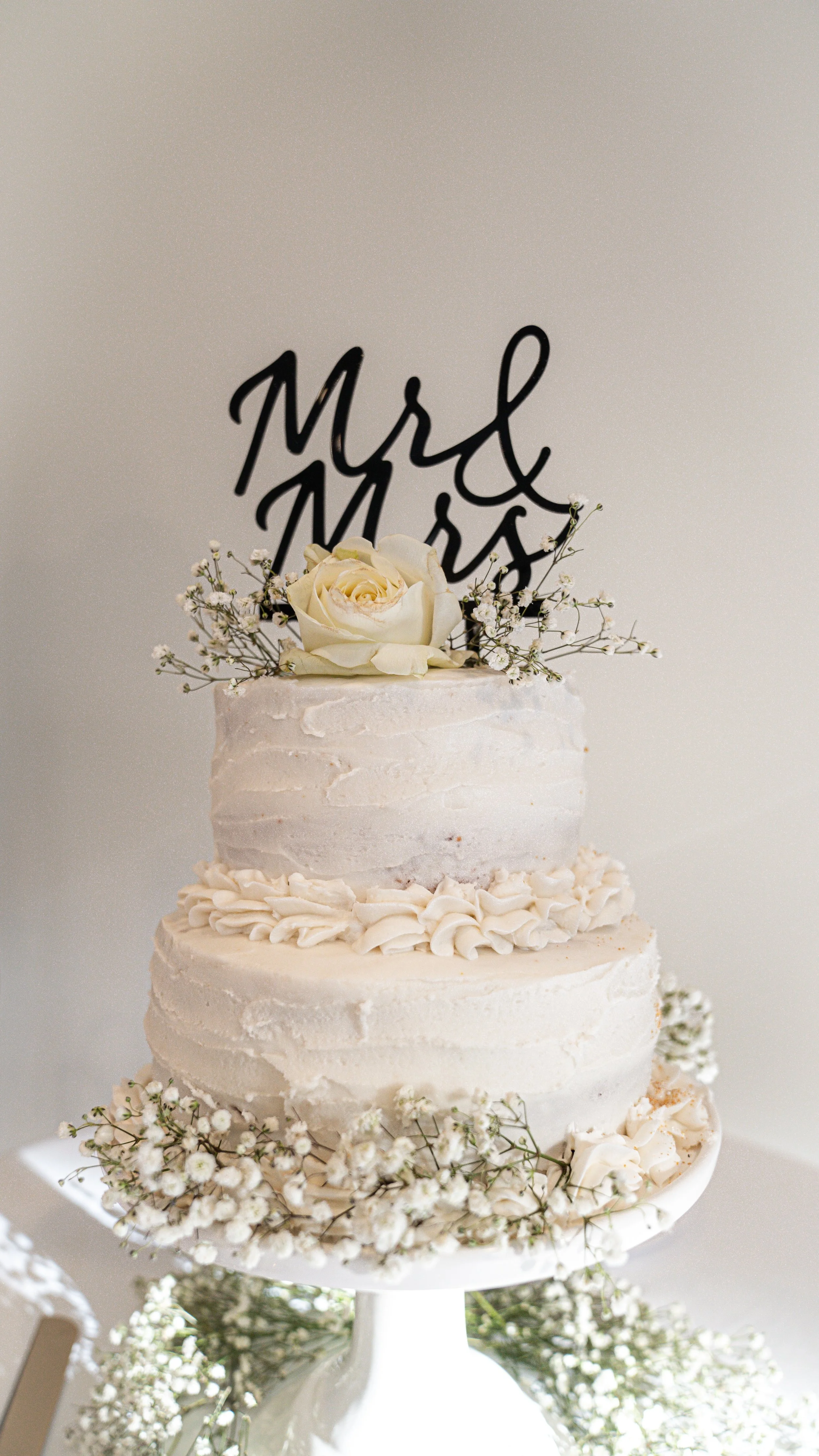 A two-tiered wedding cake decorated with white frosting, flowers, and a black cake topper that reads "mr & mrs". The cake is adorned with white roses and baby's breath, placed on a white cake stand covered with baby's breath.