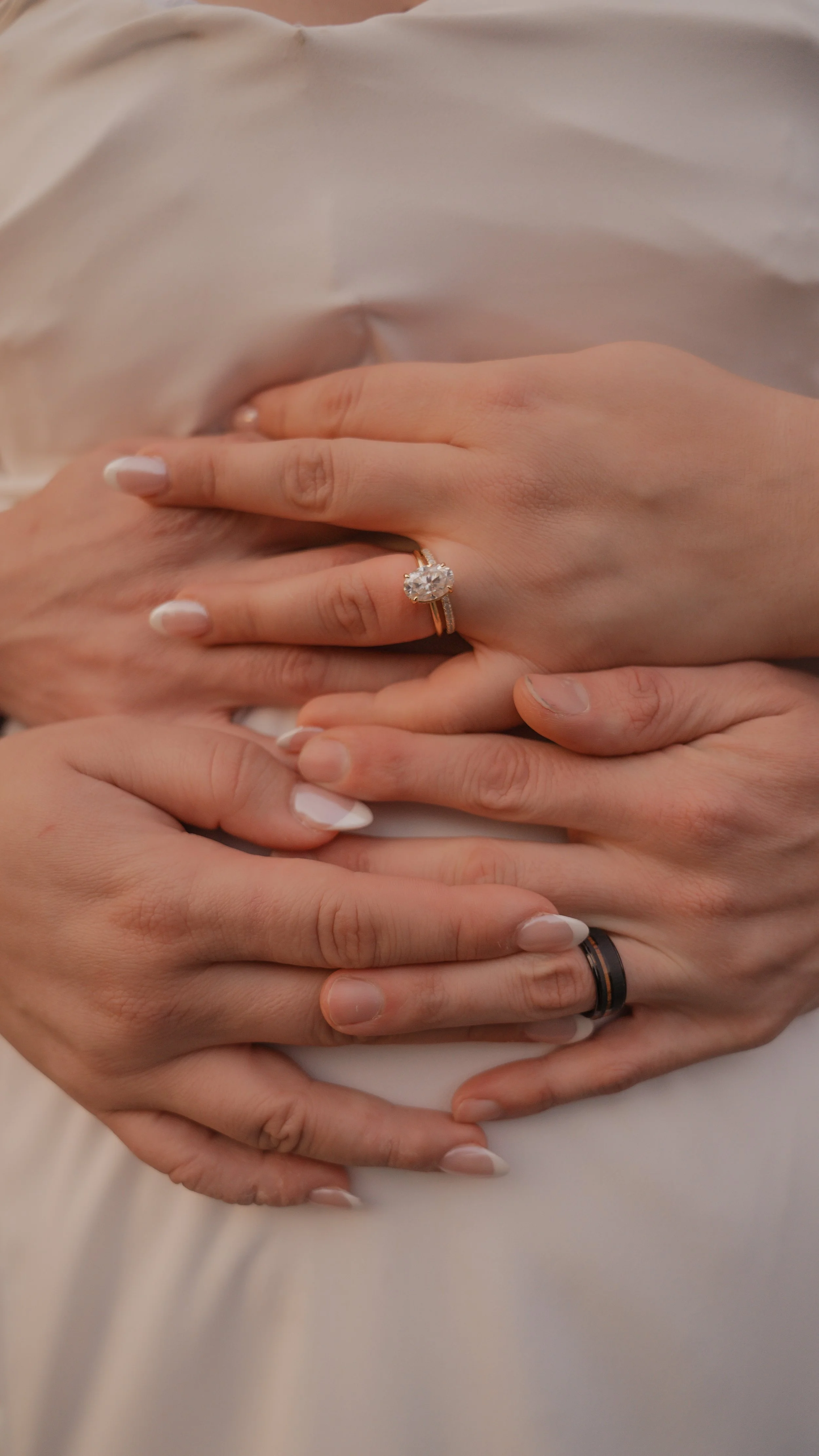Close-up of two hands with wedding rings, one with a large diamond engagement ring and the other with a black band, placed on a white fabric surface.