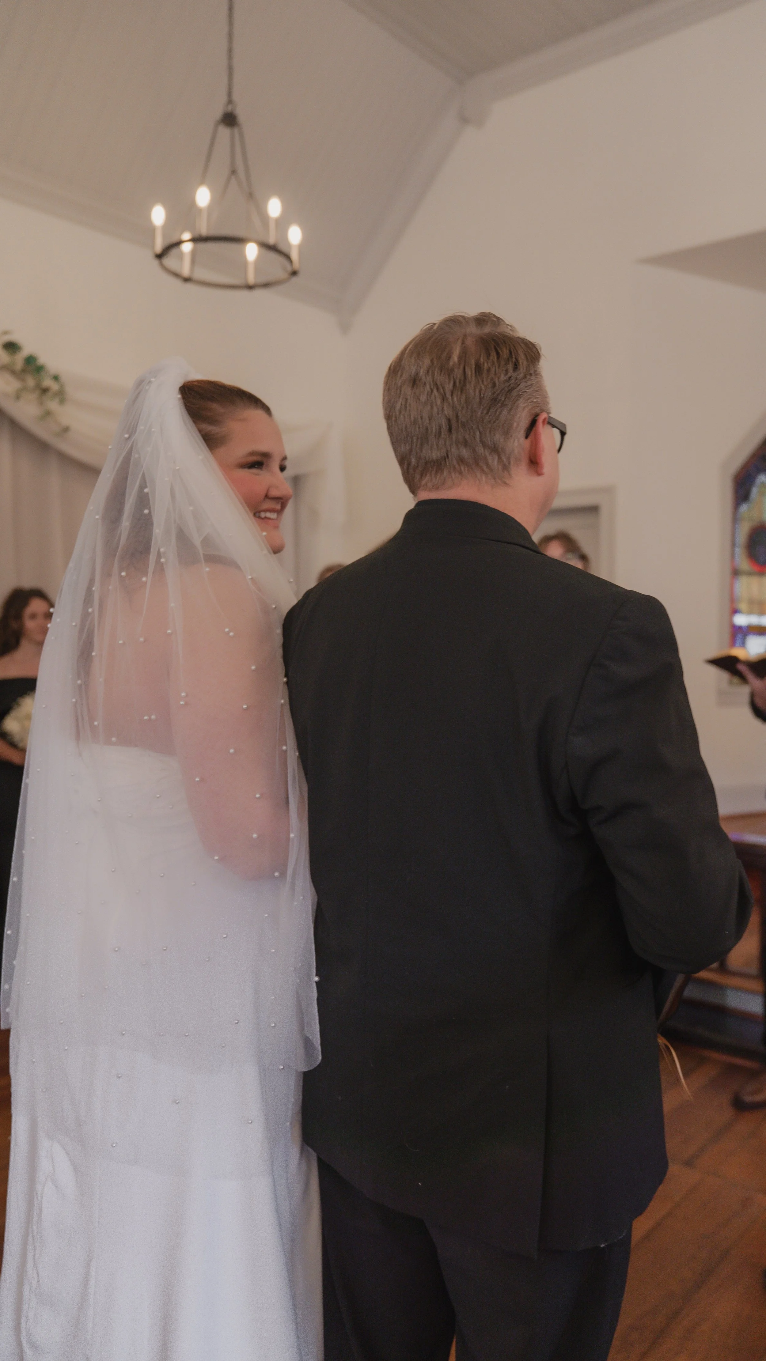 A bride smiling at her groom during a wedding ceremony indoors, with guests in the background and a stained glass window on the right.
