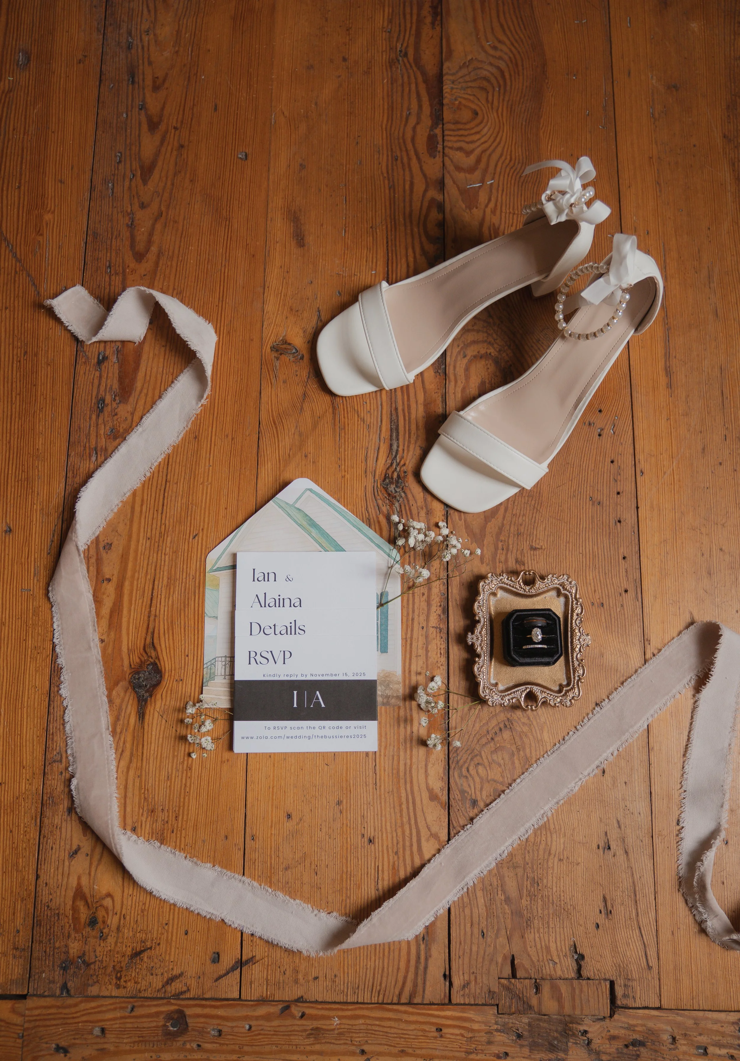 White high-heeled shoes with ankle straps, tied with white bows and pearl embellishments, placed on a wooden floor alongside wedding invitations, a vintage jewelry box with a ring, and beige fabric ribbon.