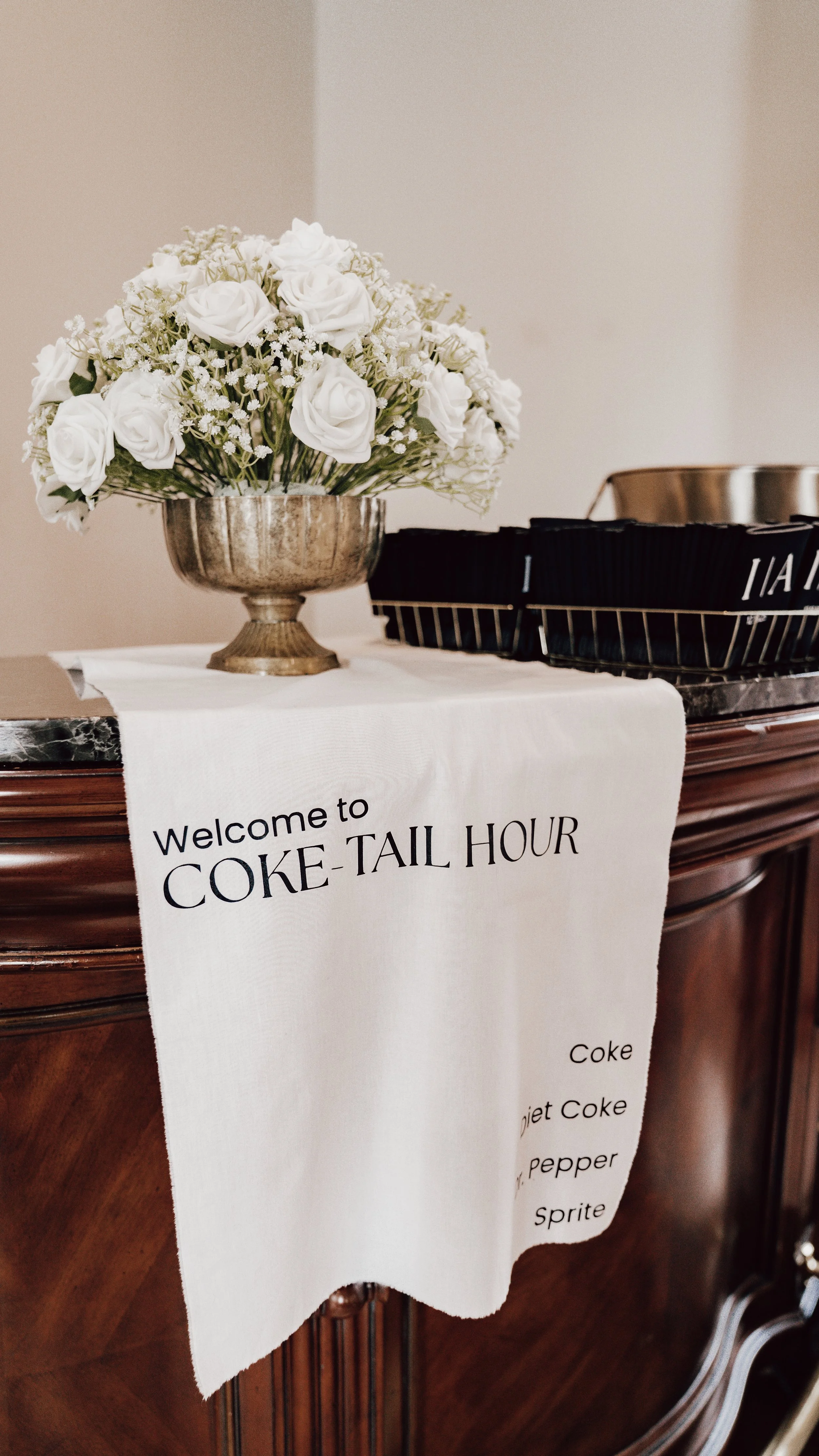 A white cloth with black text reading "Welcome to COKE TAIL HOUR" hangs over a polished wooden surface. On top, there is a silver bowl filled with white roses and small white flowers, with a black basket next to it.