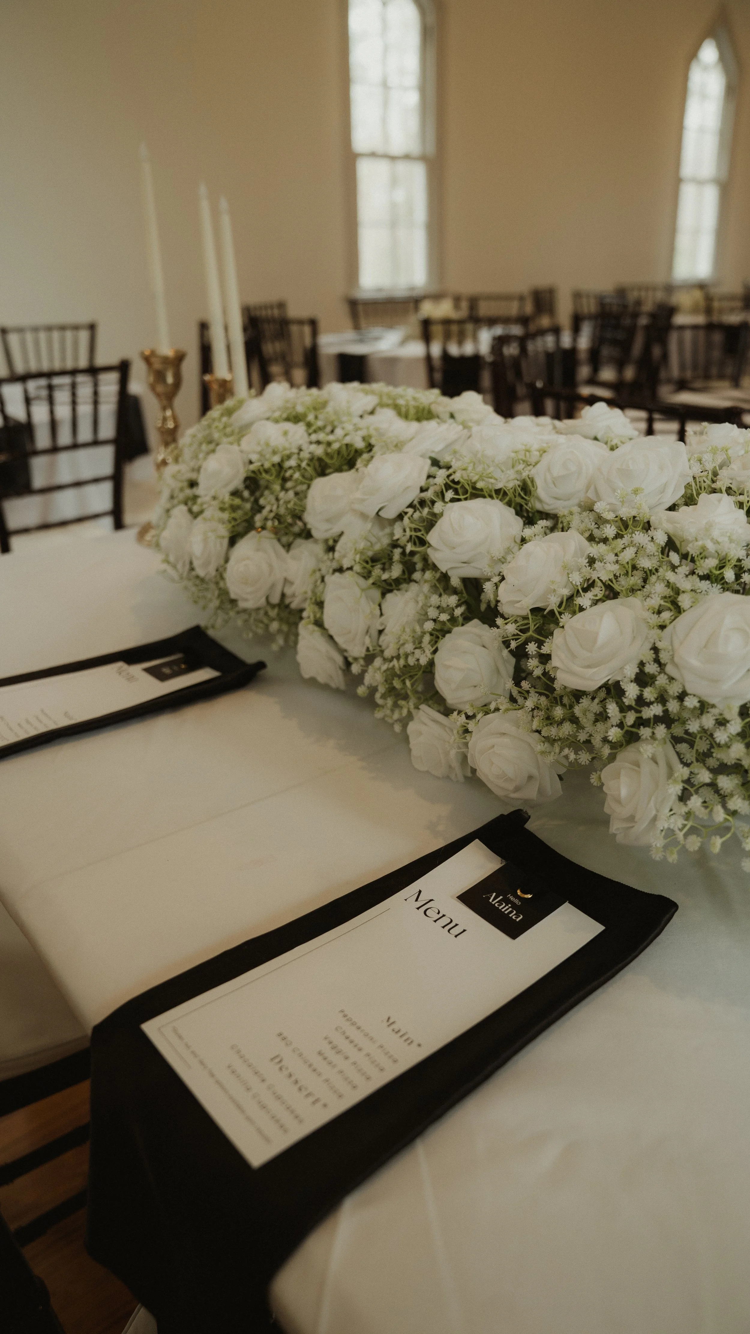 Table decorated for a formal event with a floral centerpiece of white roses and greenery, and black-covered menus.