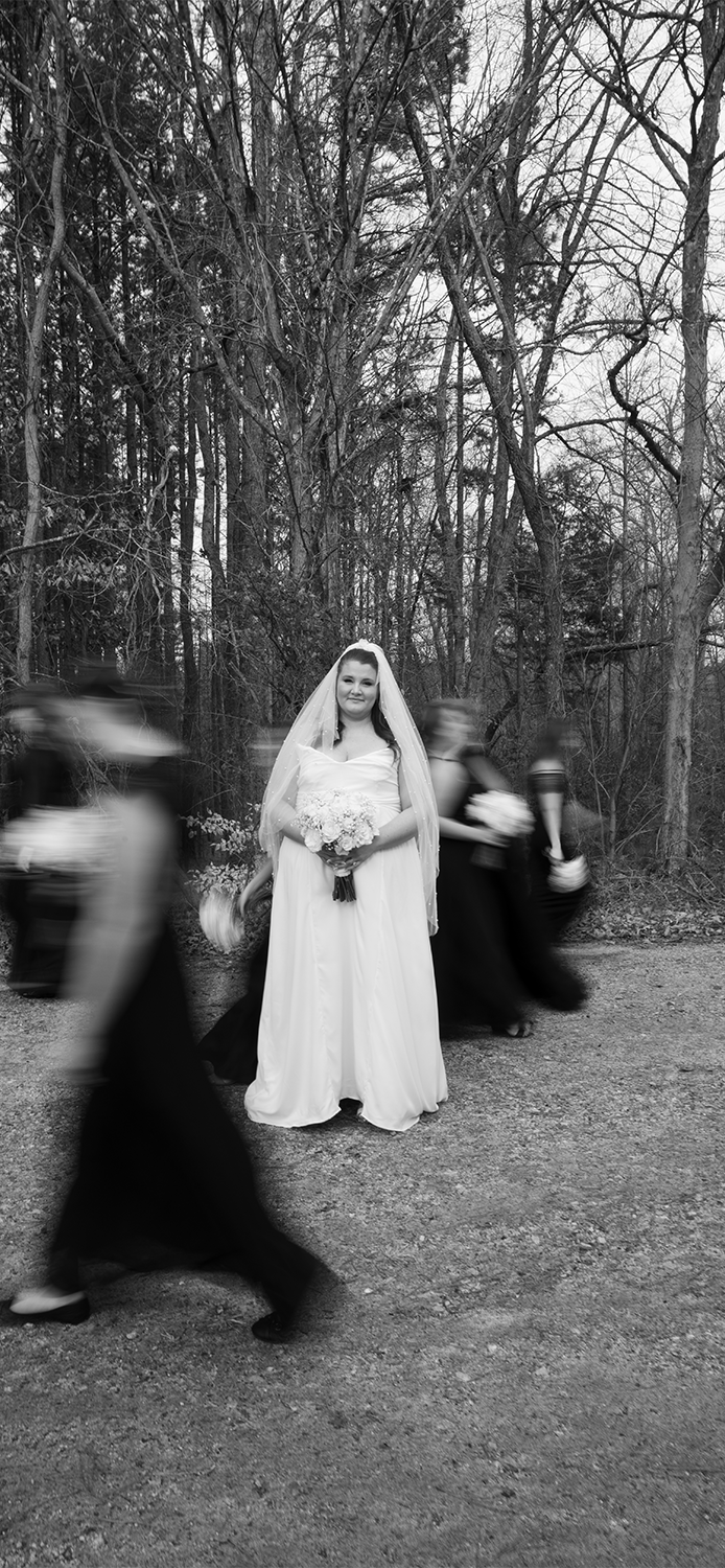 Black and white photo of a bride in a wedding gown and veil holding a bouquet, standing outdoors on a gravel path surrounded by trees, with four blurred women walking past her.
